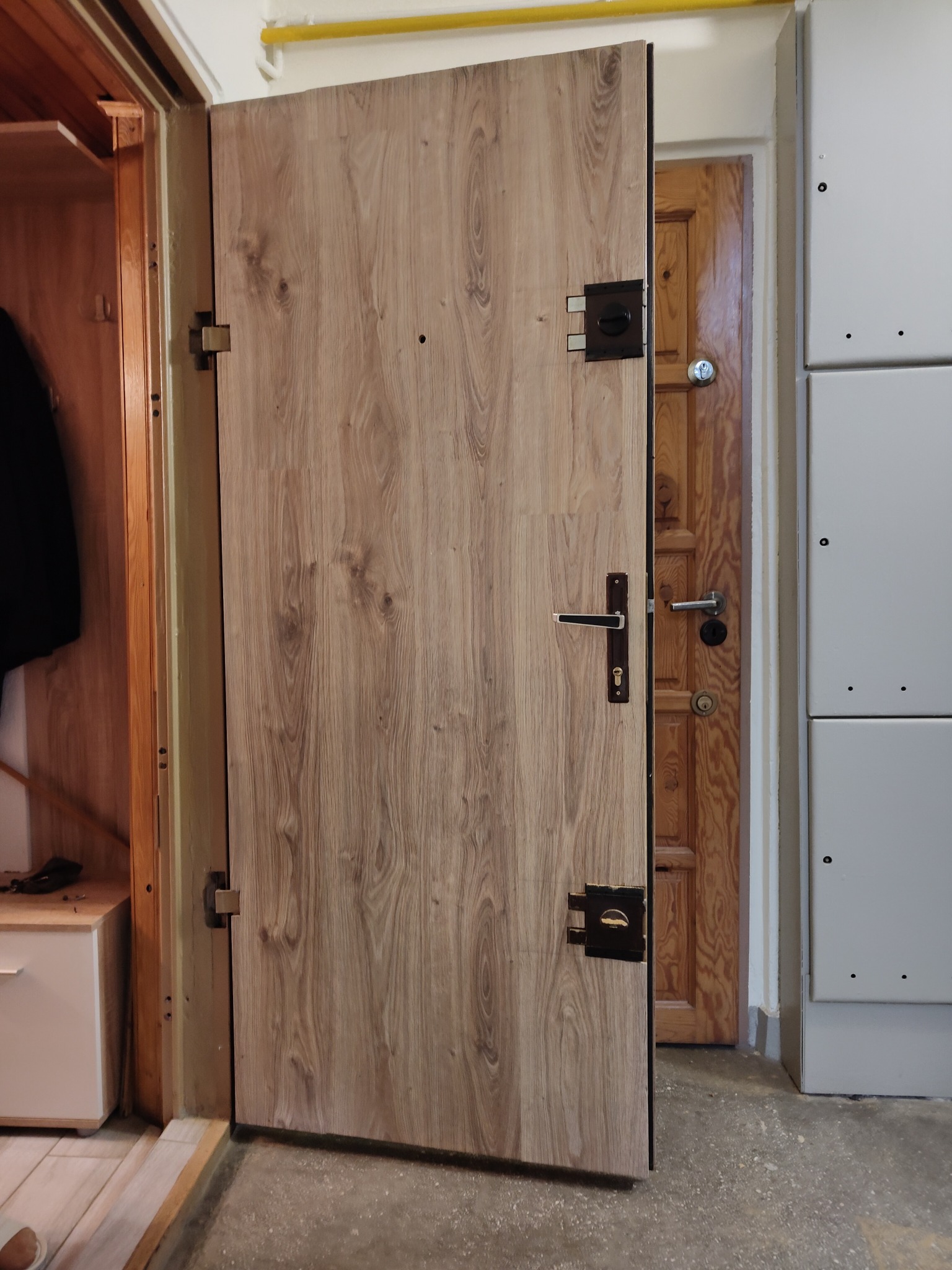 Otwarta, nowa, antywłamaniowa, drewnopodobna, brązowa okleina drzwi wejściowych z czarnymi okuciami i zamkiem, widok na drugie drzwi w tle.