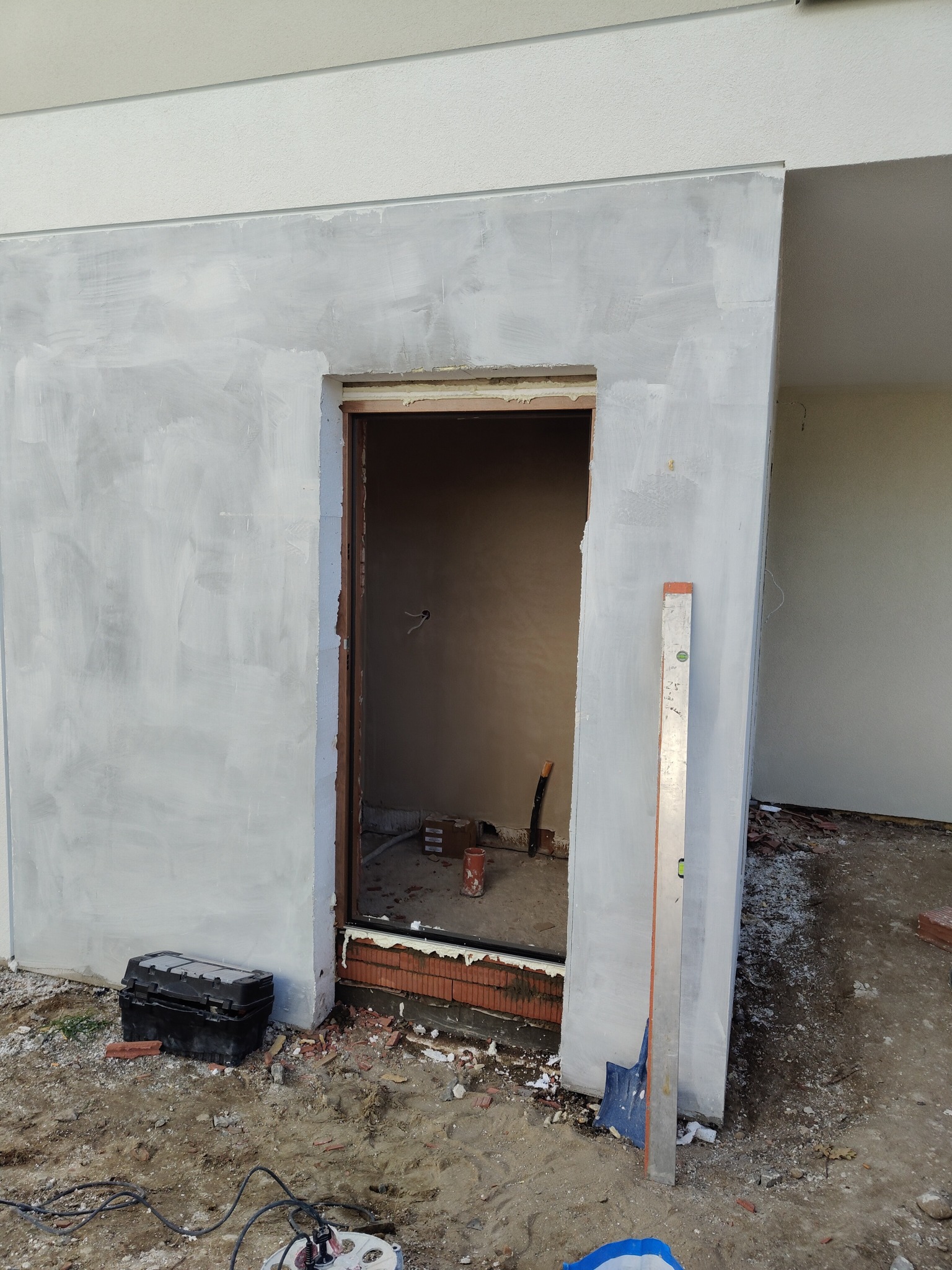 Przygotowany otwór drzwiowy w surowej ścianie, widoczny poziomica oparta o ścianę, narzędzia i gruz na ziemi.