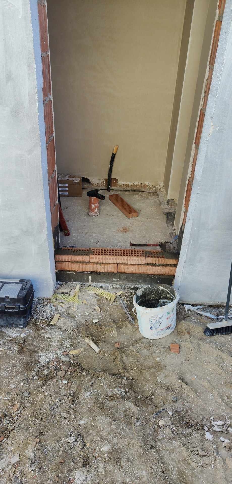 Przygotowanie do osadzania progu drzwiowego z cegieł klinkierowych, widoczny kubeł z zaprawą, narzędzia i pozostałości po pracach murarskich.
