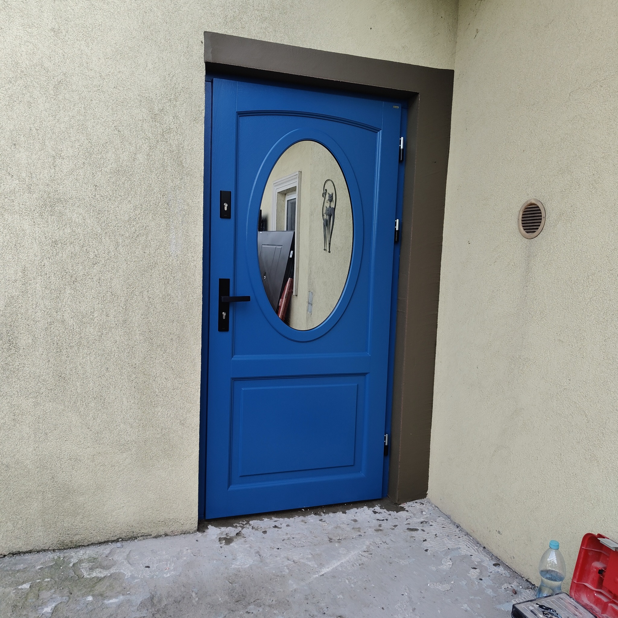 Niebieskie drzwi wejściowe z owalnym przeszkleniem, widoczne narzędzia i butelka wody, ściana o jasnej, chropowatej fakturze.