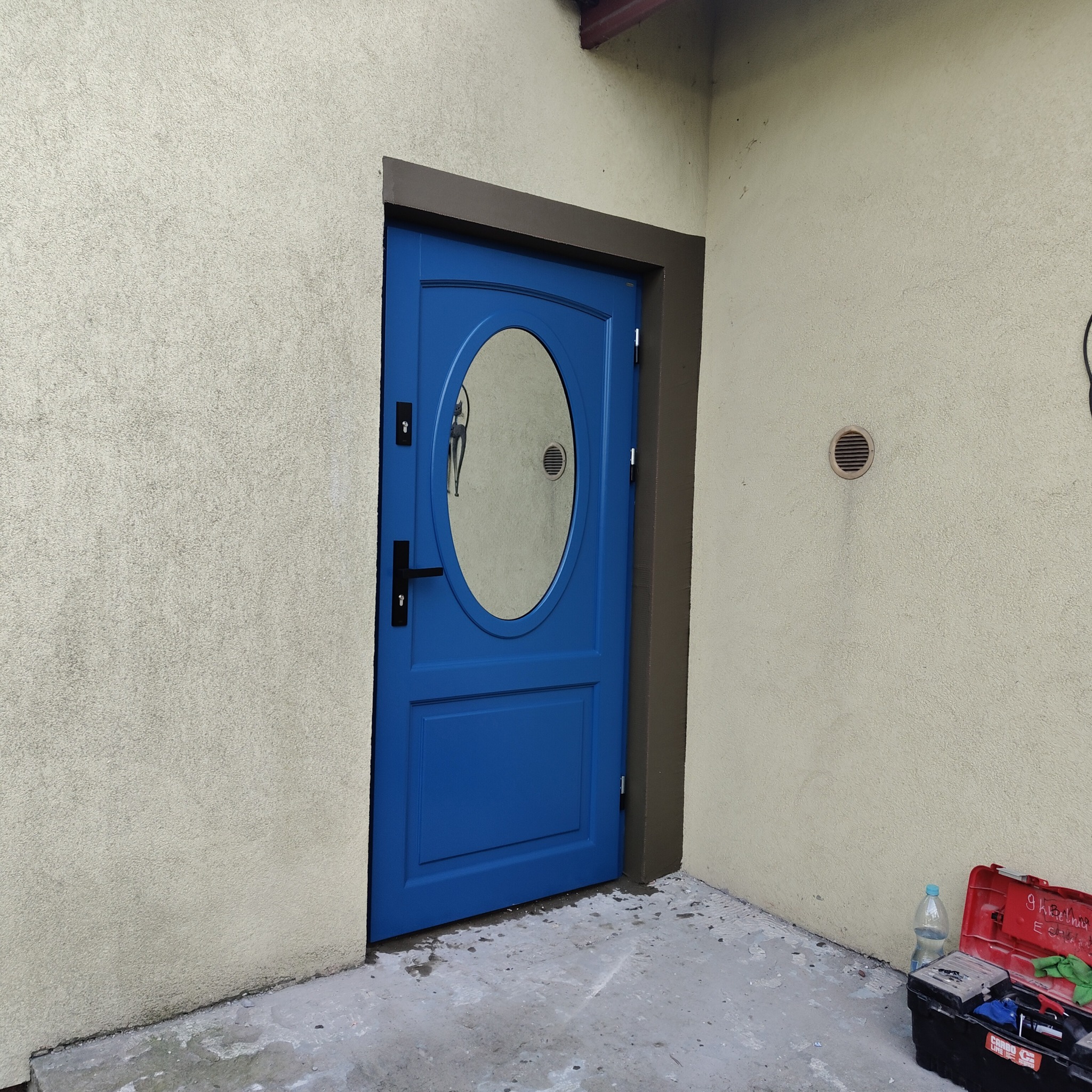 Niebieskie drzwi wejściowe z owalnym przeszkleniem i czarną klamką, zamontowane w ościeżnicy w budynku o beżowej elewacji, widoczna skrzynka z narzędziami.