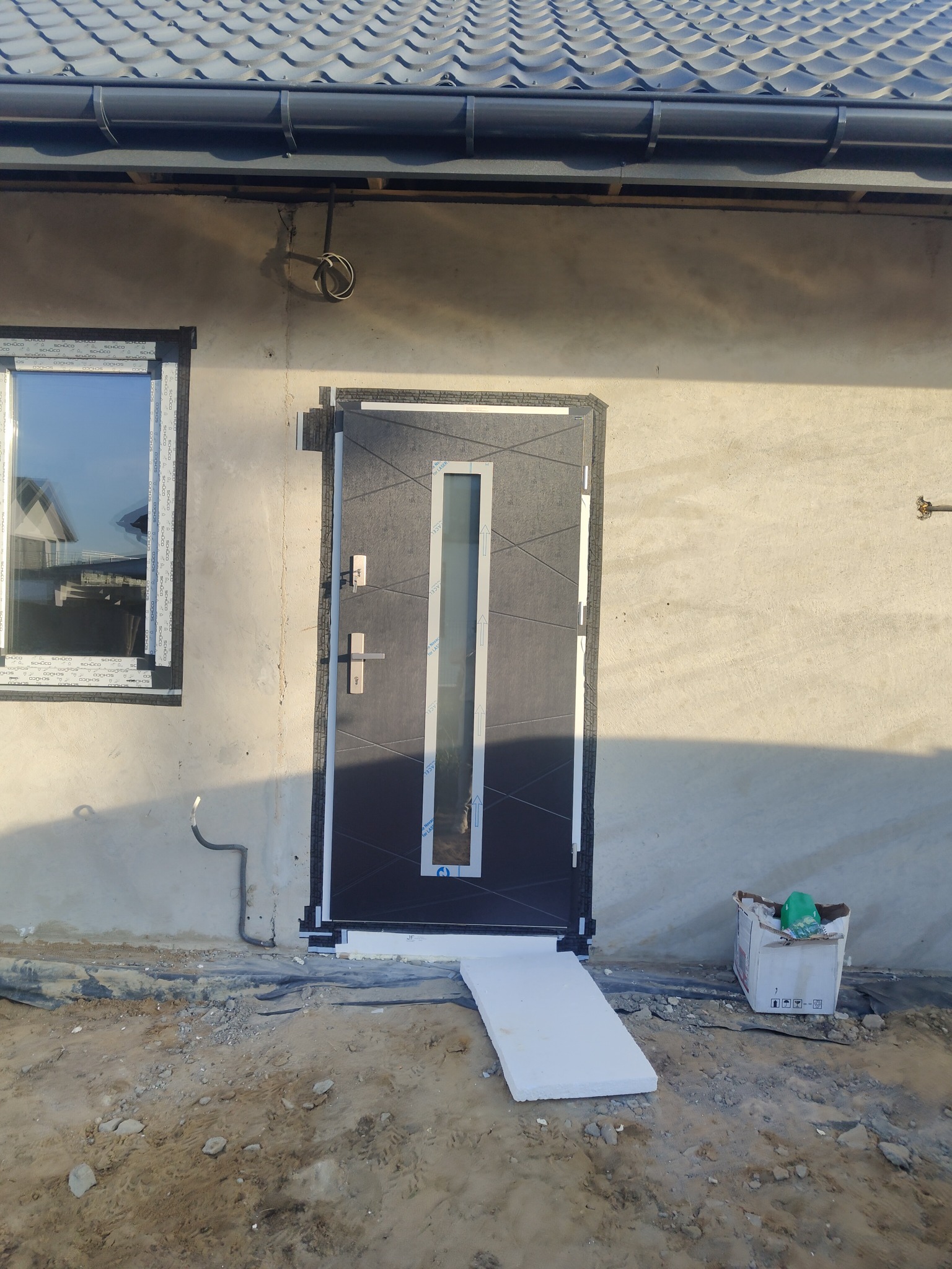 Nowe, grafitowe drzwi wejściowe z pionowym przeszkleniem, zamontowane w surowym stanie budynku, widoczne pianki montażowe i elementy ocieplenia wokół ościeżnicy, okno z naklejkami producenta obok.