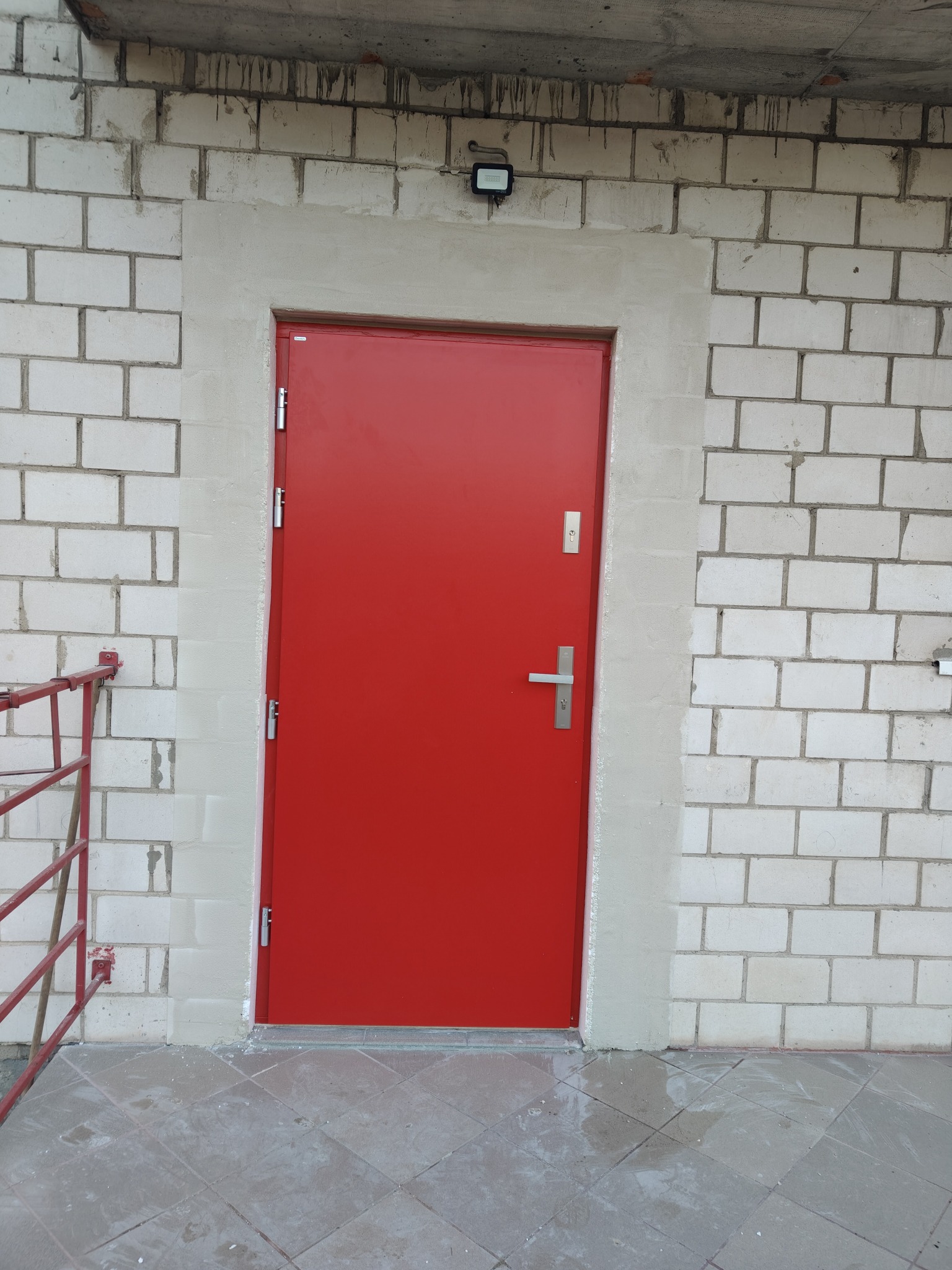Czerwone, stalowe drzwi wejściowe w surowym stanie, osadzone w ościeżnicy z szarego betonu na tle białej ściany z bloczków betonowych, widoczna lampa nad drzwiami i fragment czerwonej barierki.