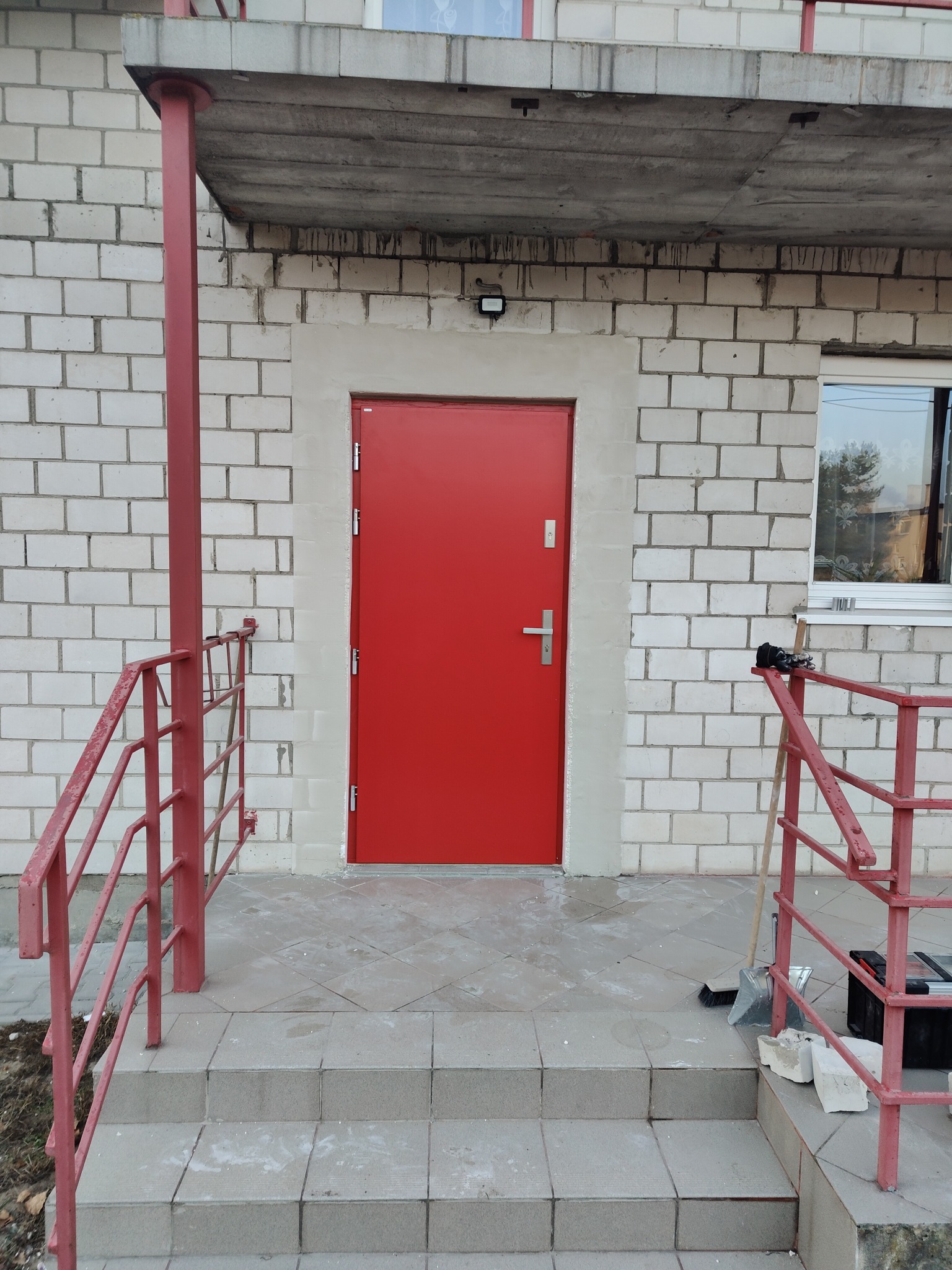 Czerwone, stalowe drzwi wejściowe do budynku z białej cegły, widoczne z zewnątrz, z metalową poręczą w kolorze czerwonym i narzędziami na schodach.