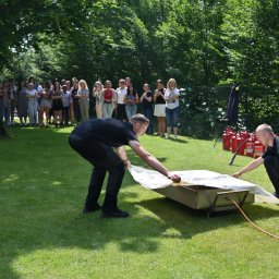 STRAŻACY - RATOWNICTWO MEDYCZNE I USŁUGI PPOŻ DAWID SZCZEPANIAK - Instruktorzy demonstrują gaszenie pożaru w metalowej wannie przy użyciu gaśnic, grupa obserwatorów w tle, trawnik w słoneczny dzień.