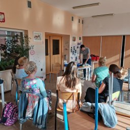 STRAŻACY - RATOWNICTWO MEDYCZNE I USŁUGI PPOŻ DAWID SZCZEPANIAK - Grupa osób uczestniczy w szkoleniu z pierwszej pomocy, instruktor demonstruje techniki na stole z rekwizytami, w tle widoczna tablica z ilustracjami i godło Polski.