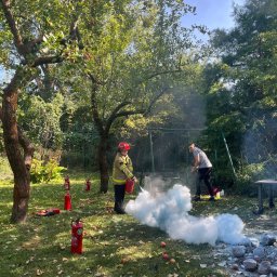 STRAŻACY - RATOWNICTWO MEDYCZNE I USŁUGI PPOŻ DAWID SZCZEPANIAK - Osoba w stroju strażaka demonstruje użycie gaśnicy na trawniku z jabłkami, w tle huśtawka i druga osoba obserwująca.