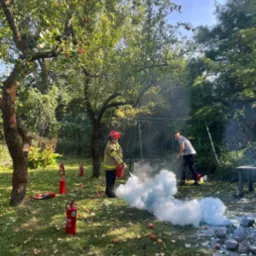 Osoba w stroju strażaka demonstruje użycie gaśnicy na trawniku z jabłkami, w tle huśtawka i druga osoba obserwująca.