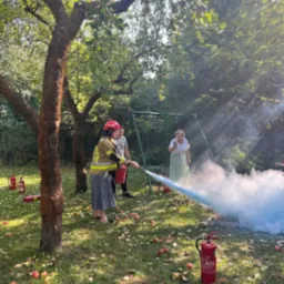 Demonstracja użycia gaśnicy proszkowej na trawniku z jabłkami, osoba w hełmie i ubraniu ochronnym kieruje strumień środka gaśniczego w stronę płomieni symulowanych w metalowej skrzynce, w tle...