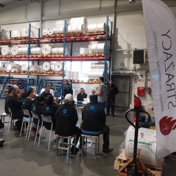 STRAŻACY - RATOWNICTWO MEDYCZNE I USŁUGI PPOŻ DAWID SZCZEPANIAK - Grupa osób w odzieży roboczej z logo KRUS siedzi na krzesłach w hali magazynowej podczas szkolenia, z widocznym banerem reklamowym firmy.