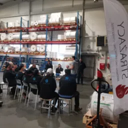 Grupa osób w odzieży roboczej z logo KRUS siedzi na krzesłach w hali magazynowej podczas szkolenia, z widocznym banerem reklamowym firmy.