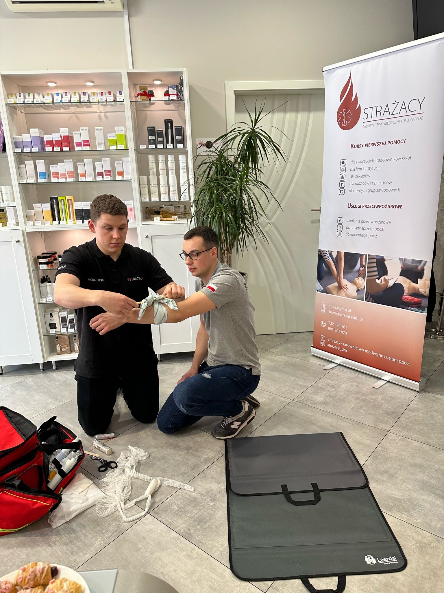 Instruktor w czarnej koszulce z logo Strażacy demonstruje technikę bandażowania przedramienia uczestnikowi kursu pierwszej pomocy, w tle regał z kosmetykami i baner reklamowy z ofertą kursów.