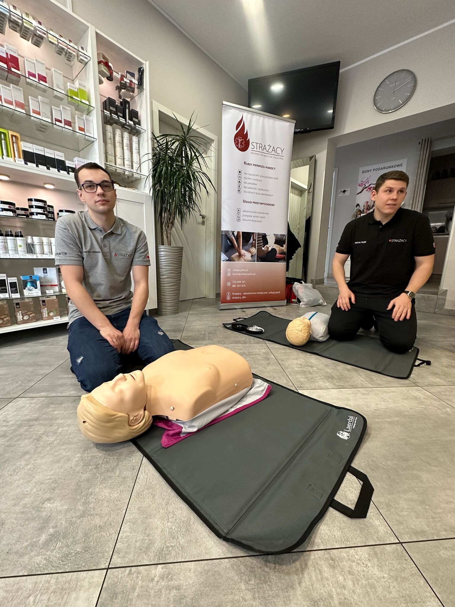 Instruktorzy klęczą obok manekinów do resuscytacji na szarych matach treningowych. W tle widoczny baner z logo i informacjami o kursach pierwszej pomocy, a także półki z kosmetykami i dekoracyjna...