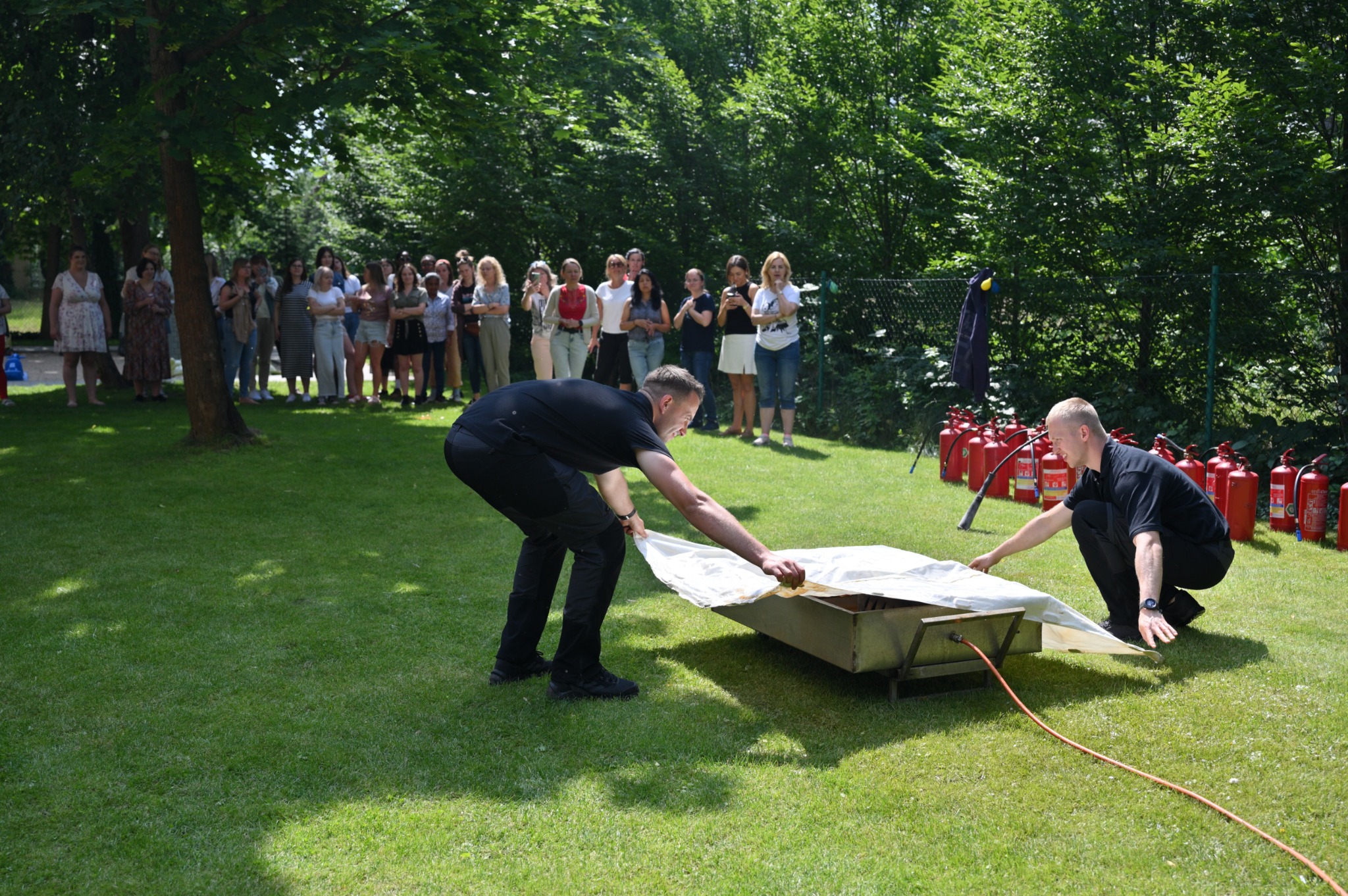 Instruktorzy demonstrują gaszenie pożaru w metalowej wannie przy użyciu gaśnic, grupa obserwatorów w tle, trawnik w słoneczny dzień.