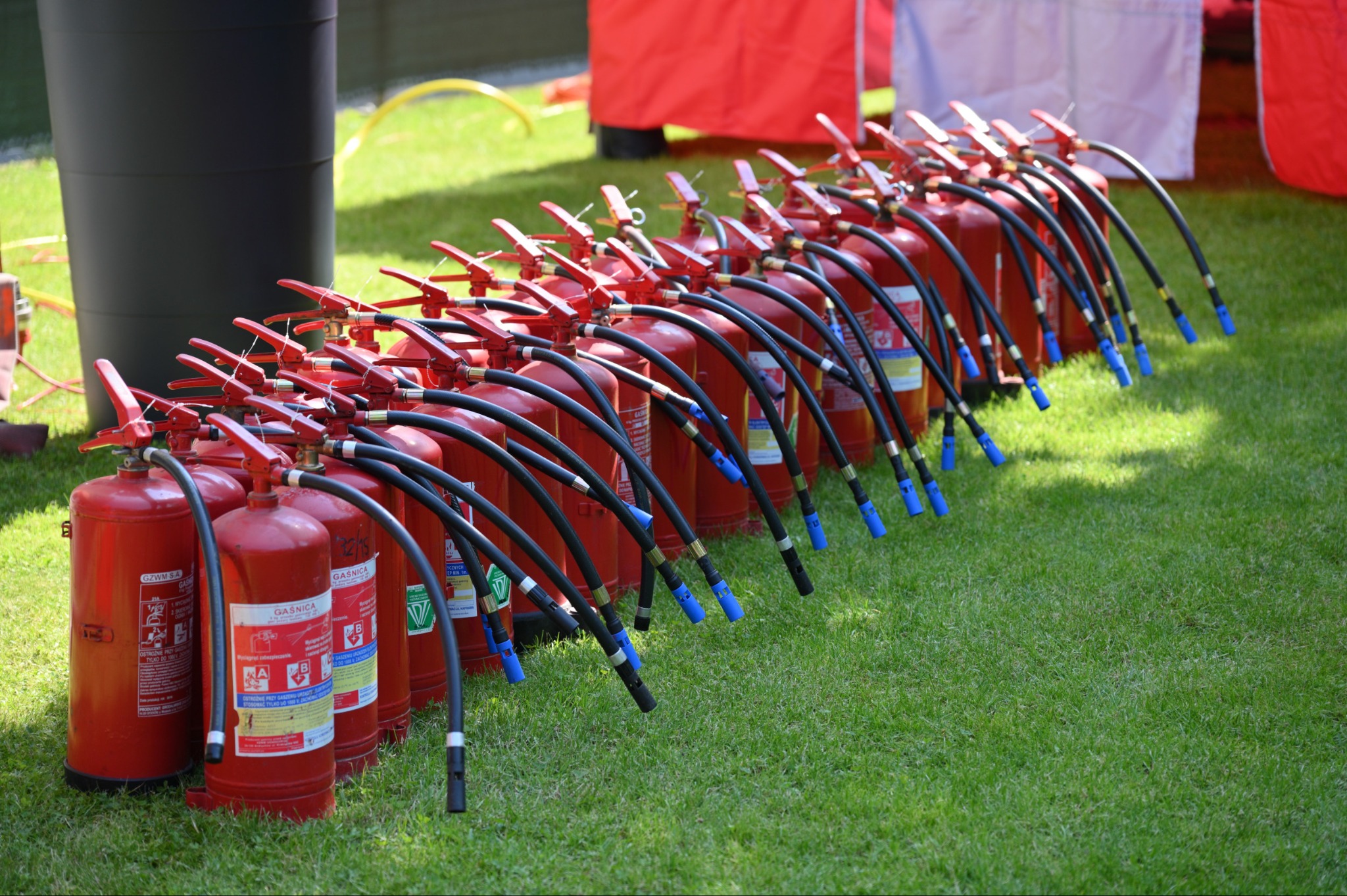 Rząd czerwonych gaśnic proszkowych z czarnymi wężami i niebieskimi końcówkami, ustawionych na trawie podczas szkolenia z zakresu bezpieczeństwa pożarowego.