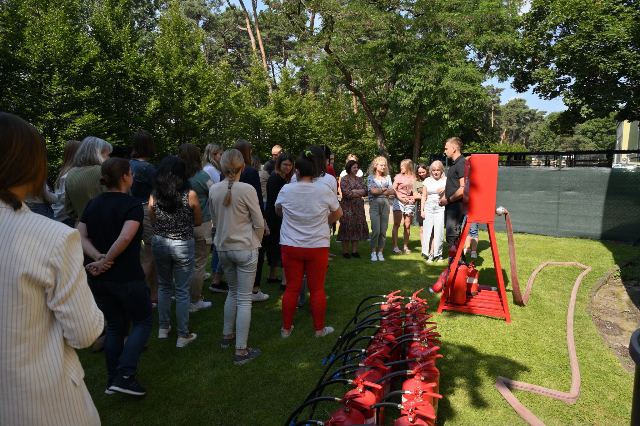 Grupa osób uczestniczy w plenerowym szkoleniu z użycia gaśnic, z rzędem czerwonych gaśnic na pierwszym planie i instruktorem objaśniającym działanie sprzętu na tle zieleni.