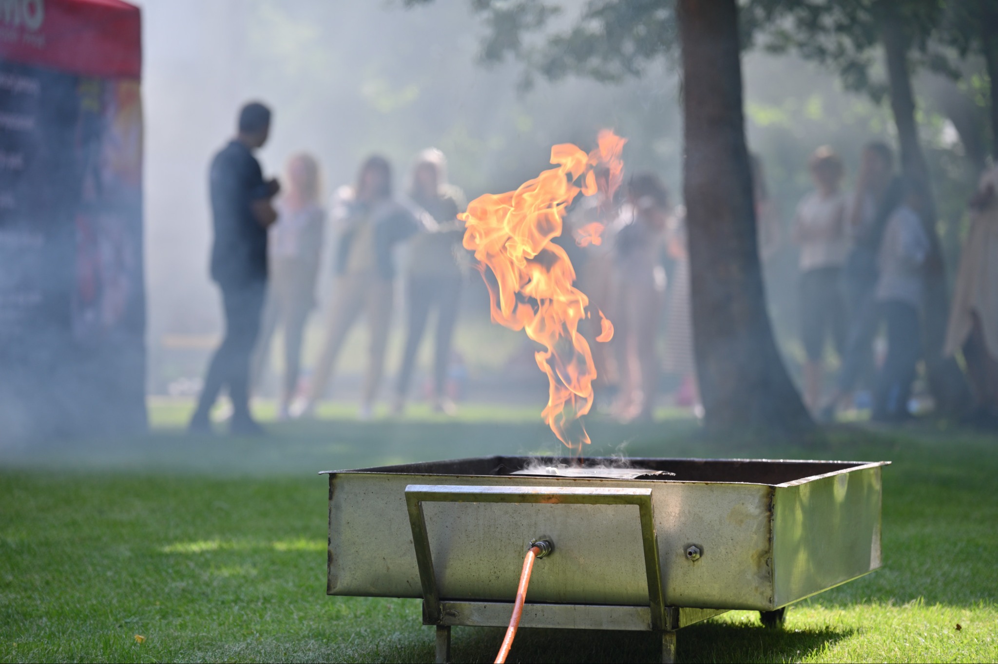 Demonstracja kontrolowanego ognia w metalowej skrzyni na trawie podczas szkolenia z zakresu bezpieczeństwa pożarowego, grupa osób w tle.