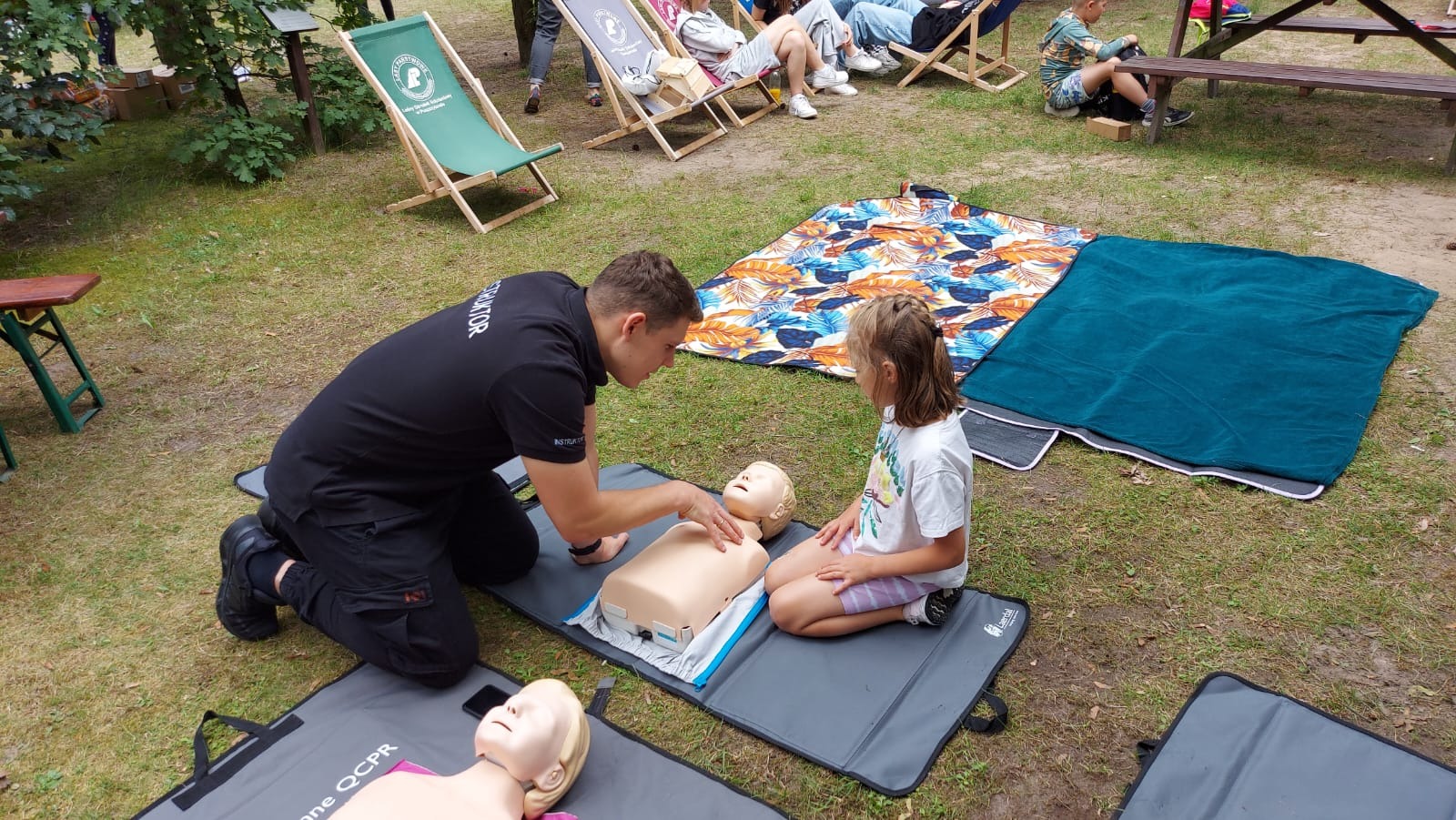 Instruktor w czarnej koszulce z napisem 'INSTRUKTOR' demonstruje resuscytację krążeniowo-oddechową na manekinie niemowlęcia, ucząc dziewczynkę siedzącą obok, w plenerze, na trawie, w tle leżaki...