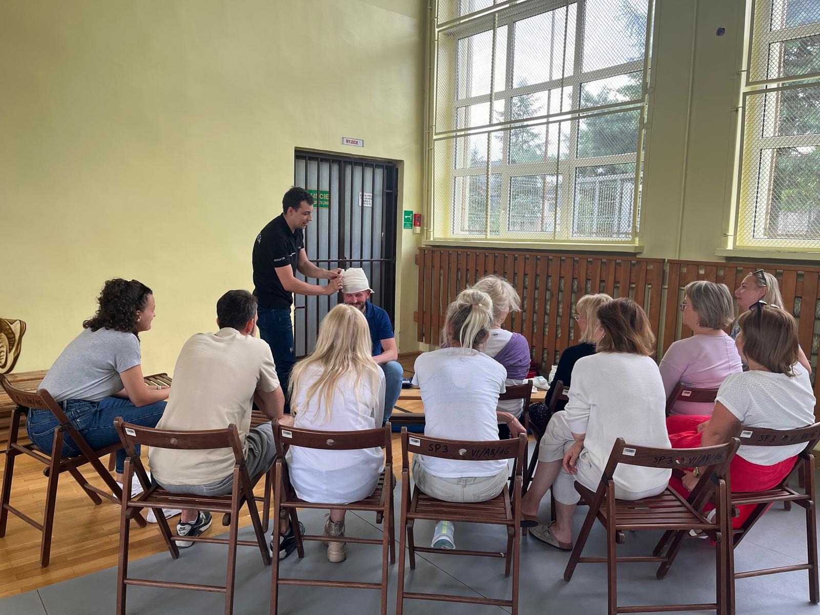 Instruktor demonstruje bandażowanie głowy na uczestniku podczas zajęć z pierwszej pomocy w sali gimnastycznej, grupa osób siedzi na krzesłach i obserwuje.