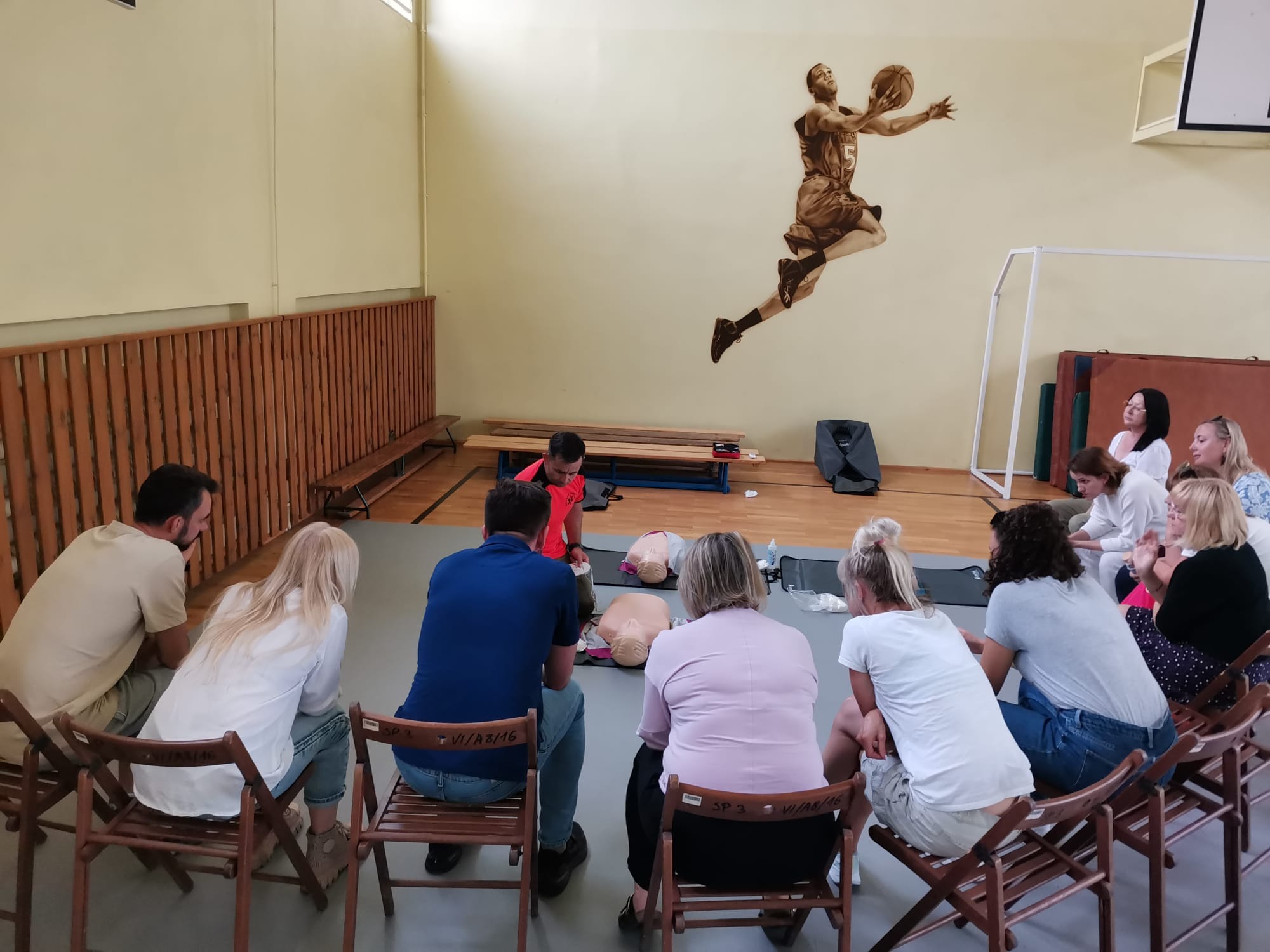 Grupa osób siedzi na krzesłach w sali gimnastycznej podczas kursu pierwszej pomocy, instruktor demonstruje resuscytację na manekinie, na ścianie widoczny mural z koszykarzem.