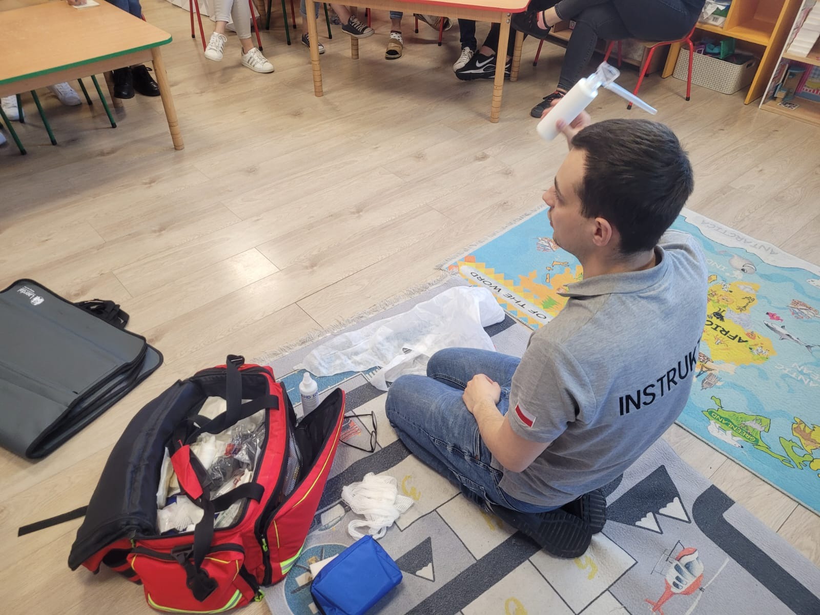 Instruktor siedzi na dywanie z mapą świata, demonstrując użycie maski resuscytacyjnej; obok rozłożona torba medyczna z wyposażeniem.