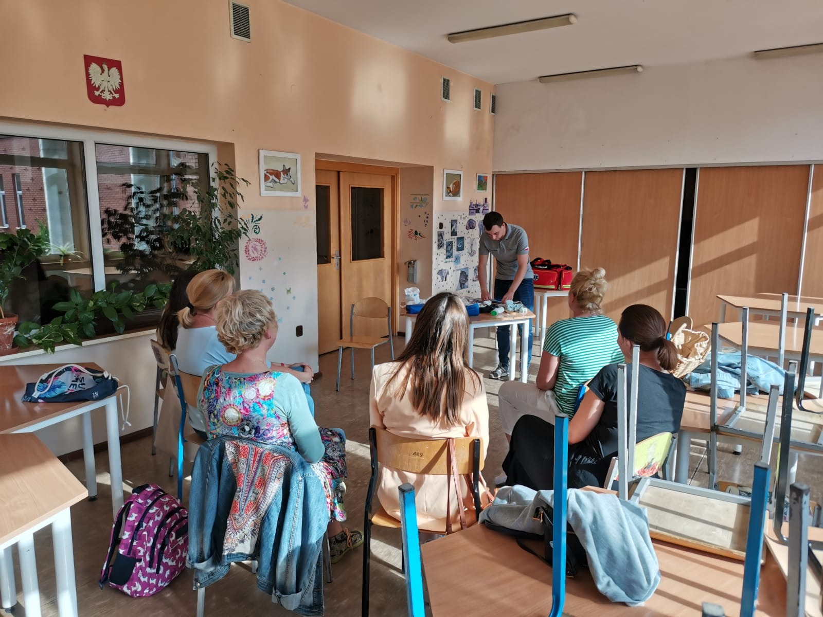 Grupa osób uczestniczy w szkoleniu z pierwszej pomocy, instruktor demonstruje techniki na stole z rekwizytami, w tle widoczna tablica z ilustracjami i godło Polski.