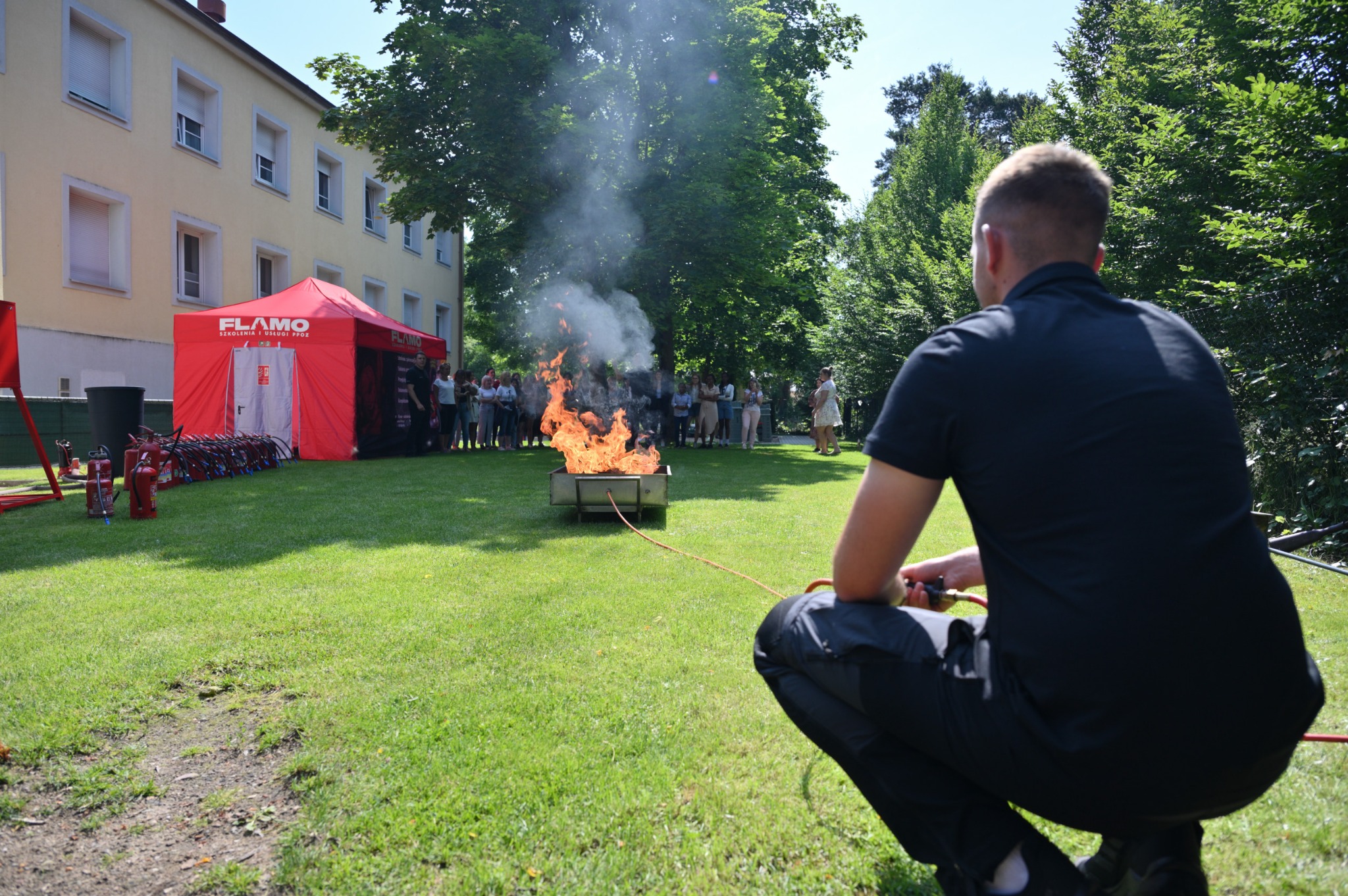 Osoba w kucki obsługuje urządzenie do gaszenia ognia w metalowej skrzyni na trawniku, w tle grupa osób, czerwony namiot z napisem FLAMO i rzędem gaśnic.