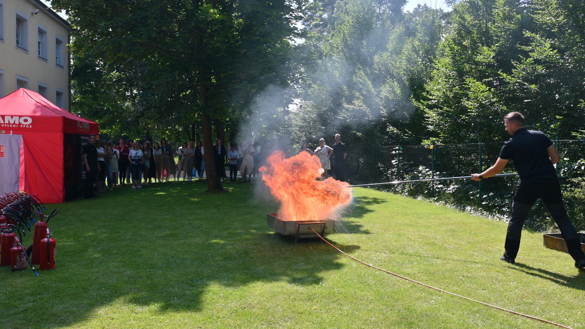 Instruktor demonstruje gaszenie płonącego ognia w metalowej skrzyni podczas pokazu dla grupy obserwatorów, z widocznymi gaśnicami w tle.