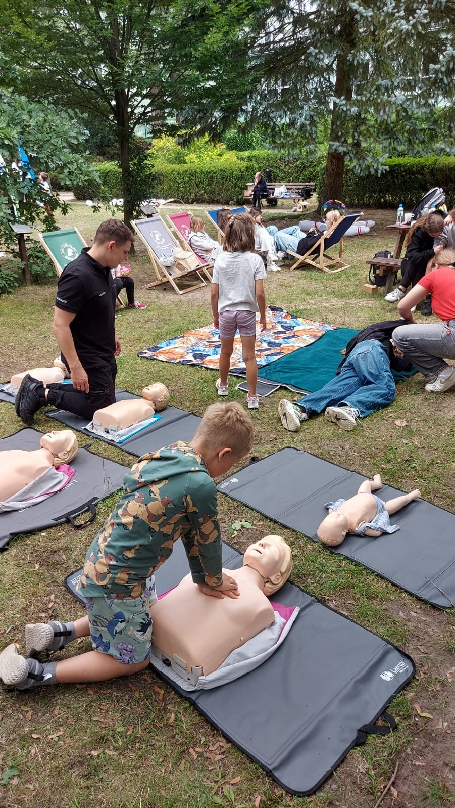 Szkolenie z pierwszej pomocy na świeżym powietrzu: instruktor demonstruje resuscytację na manekinie, dzieci ćwiczą pod nadzorem, w tle relaks na leżakach.
