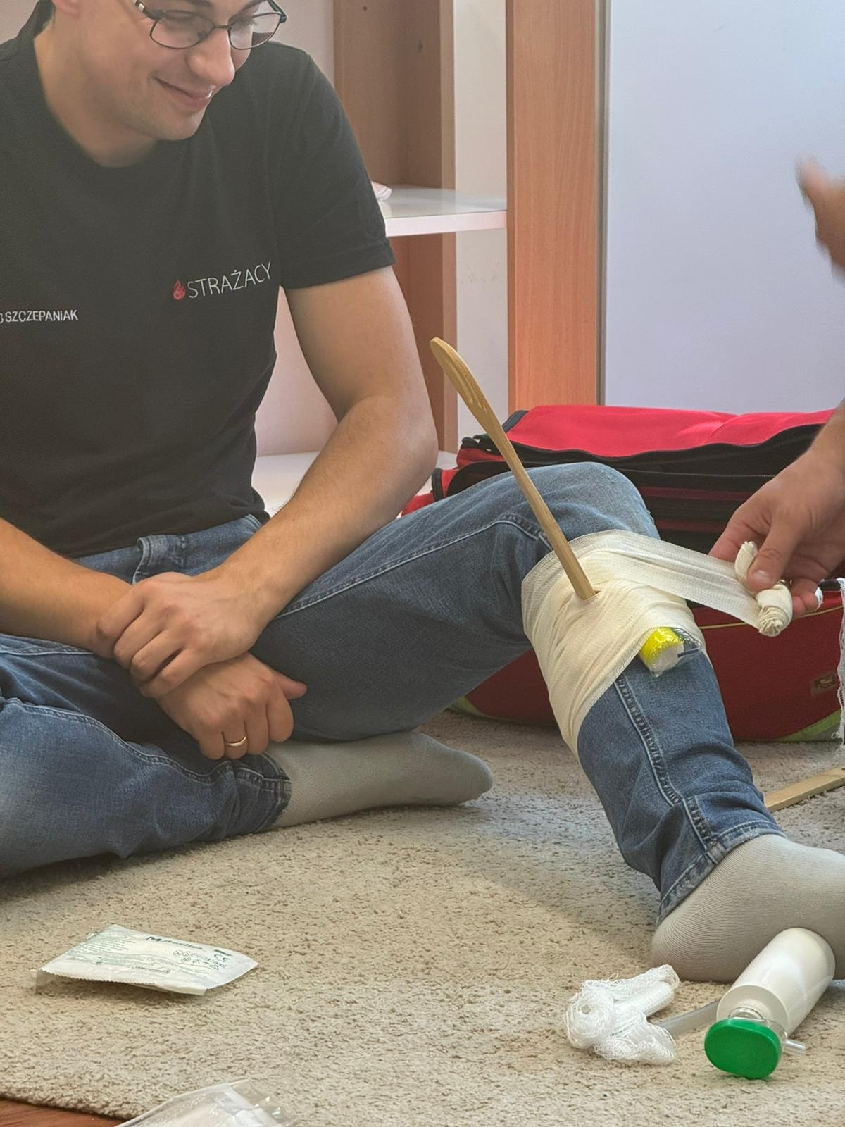 Instruktor z uśmiechem demonstruje usztywnienie złamanej nogi z użyciem drewnianej łyżki i bandaża elastycznego, na podłodze leżą materiały opatrunkowe, w tle torba medyczna i regał.
