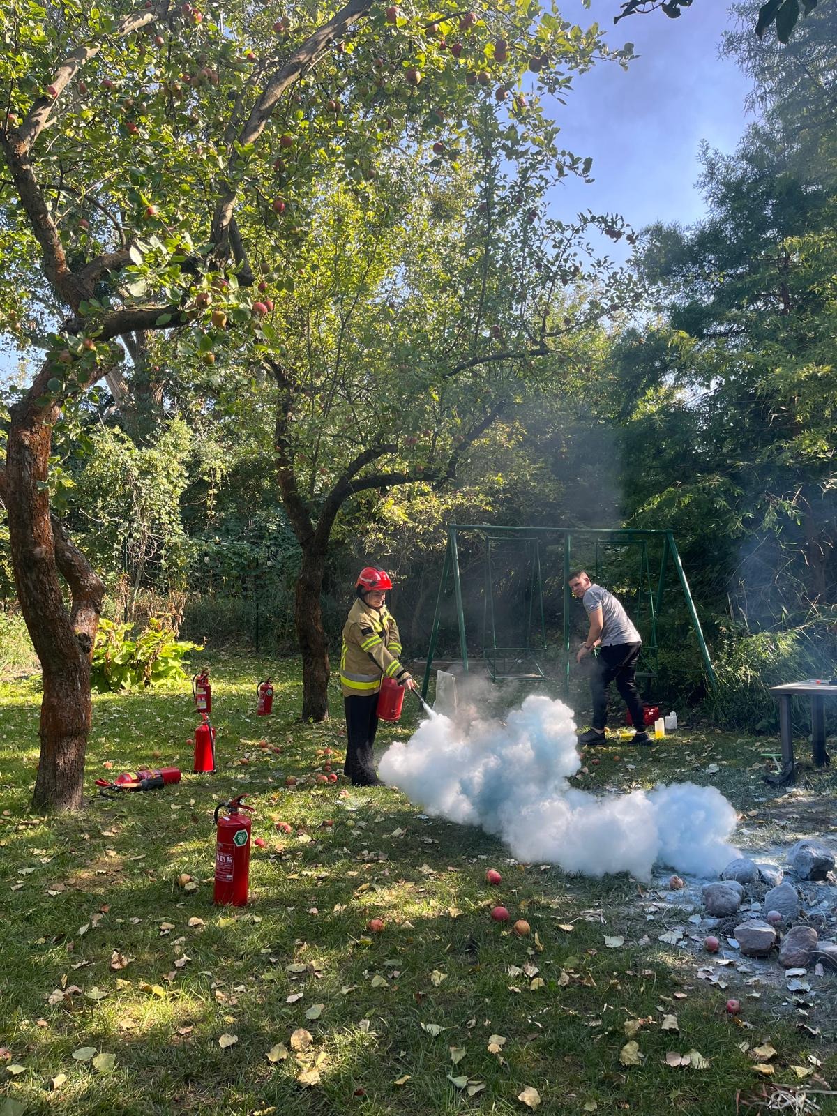 Osoba w stroju strażaka demonstruje użycie gaśnicy na trawniku z jabłkami, w tle huśtawka i druga osoba obserwująca.
