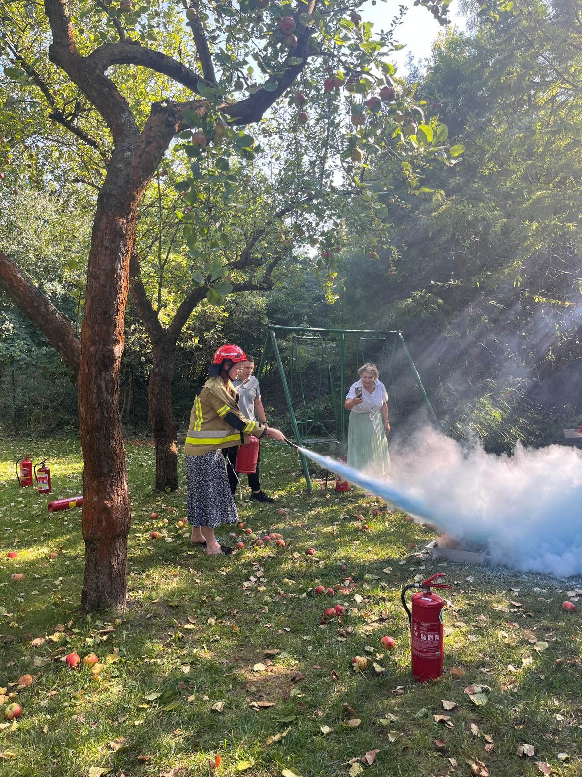 Demonstracja użycia gaśnicy proszkowej na trawniku z jabłkami, osoba w hełmie i ubraniu ochronnym kieruje strumień środka gaśniczego w stronę płomieni symulowanych w metalowej skrzynce, w tle...