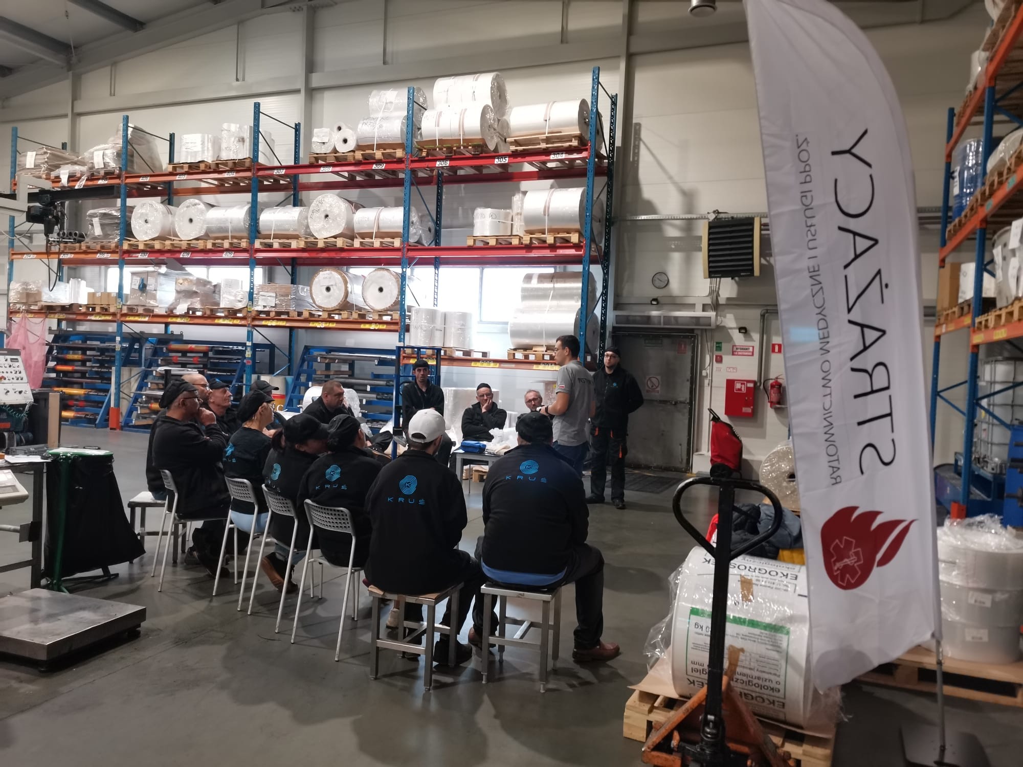 Grupa osób w odzieży roboczej z logo KRUS siedzi na krzesłach w hali magazynowej podczas szkolenia, z widocznym banerem reklamowym firmy.