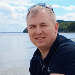 Portret uśmiechniętego mężczyzny w ciemnej koszuli polo z okularami na głowie, na tle jeziora i błękitnego nieba z chmurami.