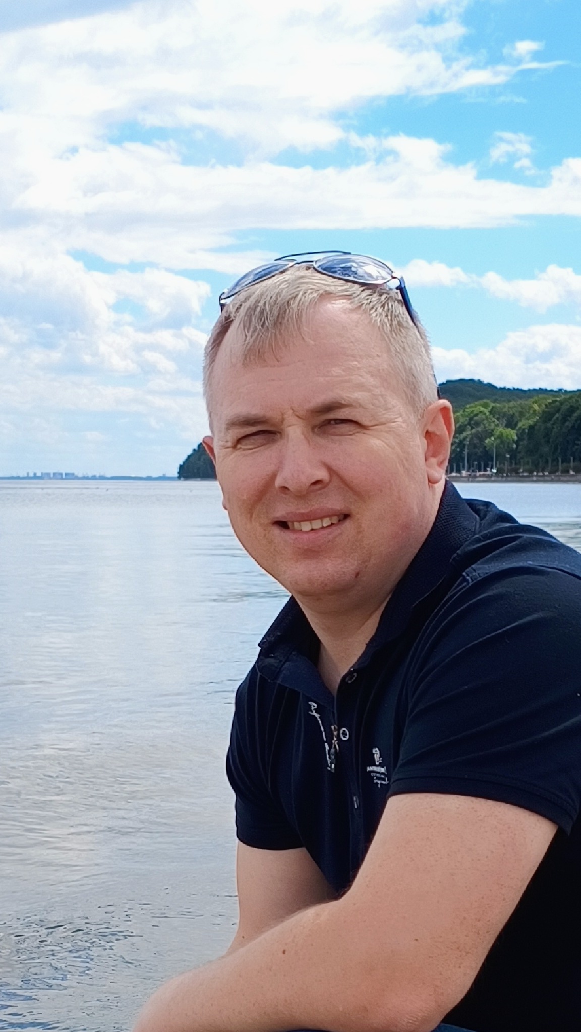 Portret uśmiechniętego mężczyzny w ciemnej koszuli polo z okularami na głowie, na tle jeziora i błękitnego nieba z chmurami.