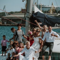Andrii Kotelnikov - Duża grupa osób, w tym panna młoda w koronkowej sukni, pozuje na jachcie w porcie, z dźwigami widocznymi w tle.