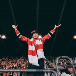 Andrii Kotelnikov - Uśmiechnięty chłopiec w czerwono-białym szaliku i czapce z daszkiem, z rozłożonymi rękami, świętuje na trybunach stadionu podczas wydarzenia sportowego, z tłumem kibiców w tle.