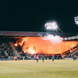 Andrii Kotelnikov - Stadion piłkarski podczas meczu, kibice odpalają race, tworząc pomarańczową zasłonę dymu na trybunach, widoczni piłkarze na boisku i tablica wyników.