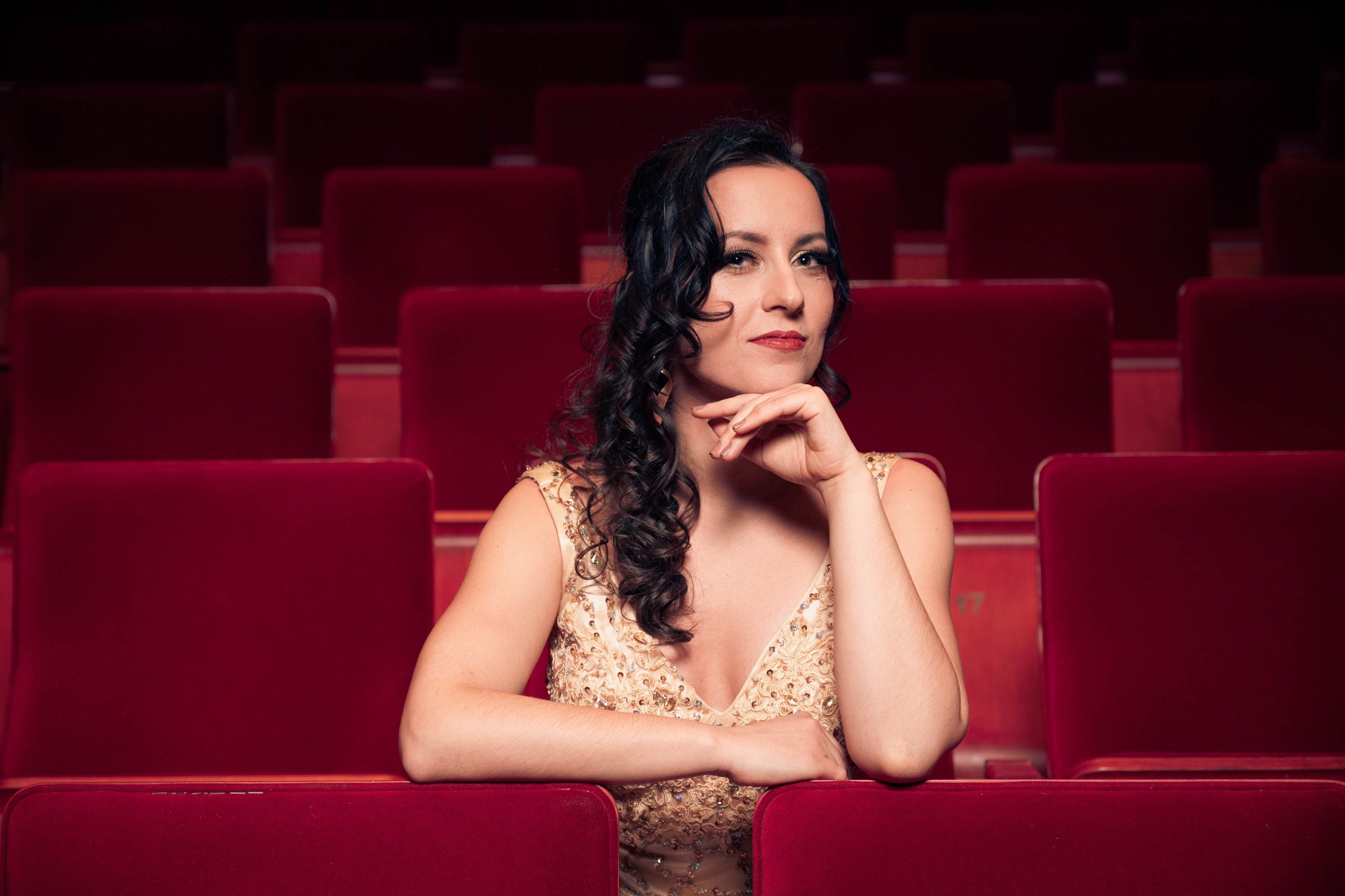 Portret kobiety w złotej sukni siedzącej w czerwonym fotelu w teatrze, z ręką podpartą pod brodą i zamyślonym wyrazem twarzy.