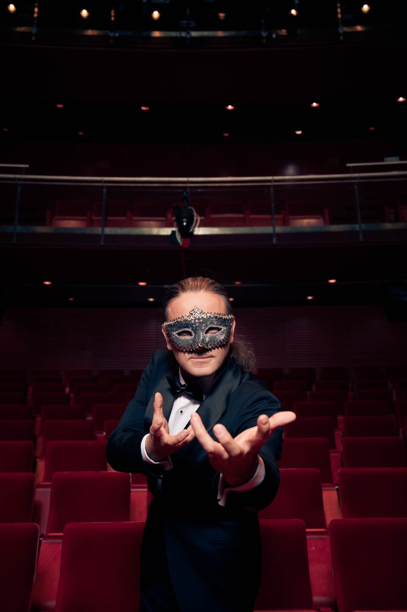 Mężczyzna w muszce i zdobionej masce, stojący na tle pustej widowni teatru, z wyciągniętymi dłońmi, jakby zapraszał na przedstawienie.
