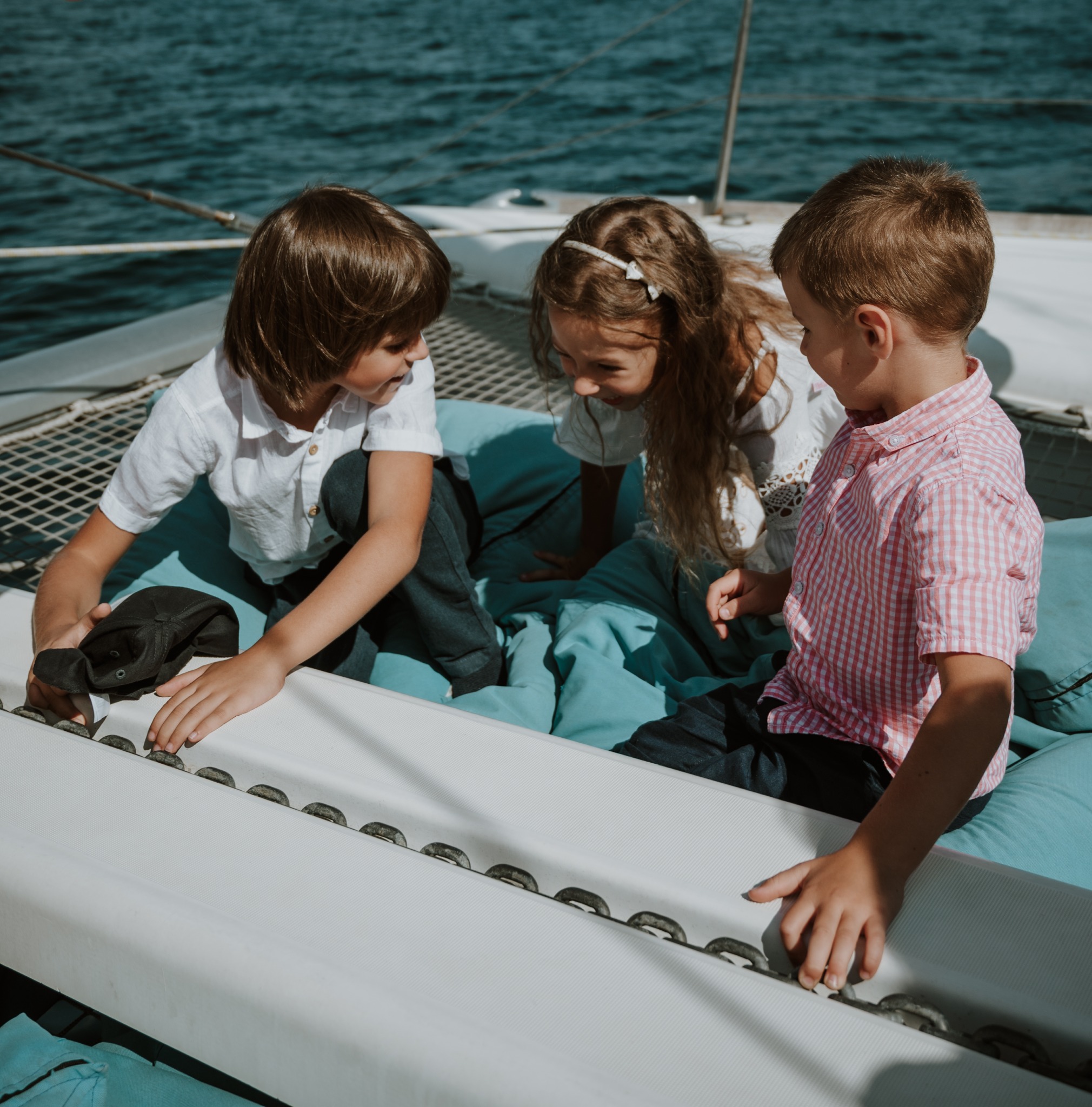 Troje dzieci siedzi na niebieskich poduszkach na pokładzie katamaranu, jedno z nich trzyma czarną czapkę
