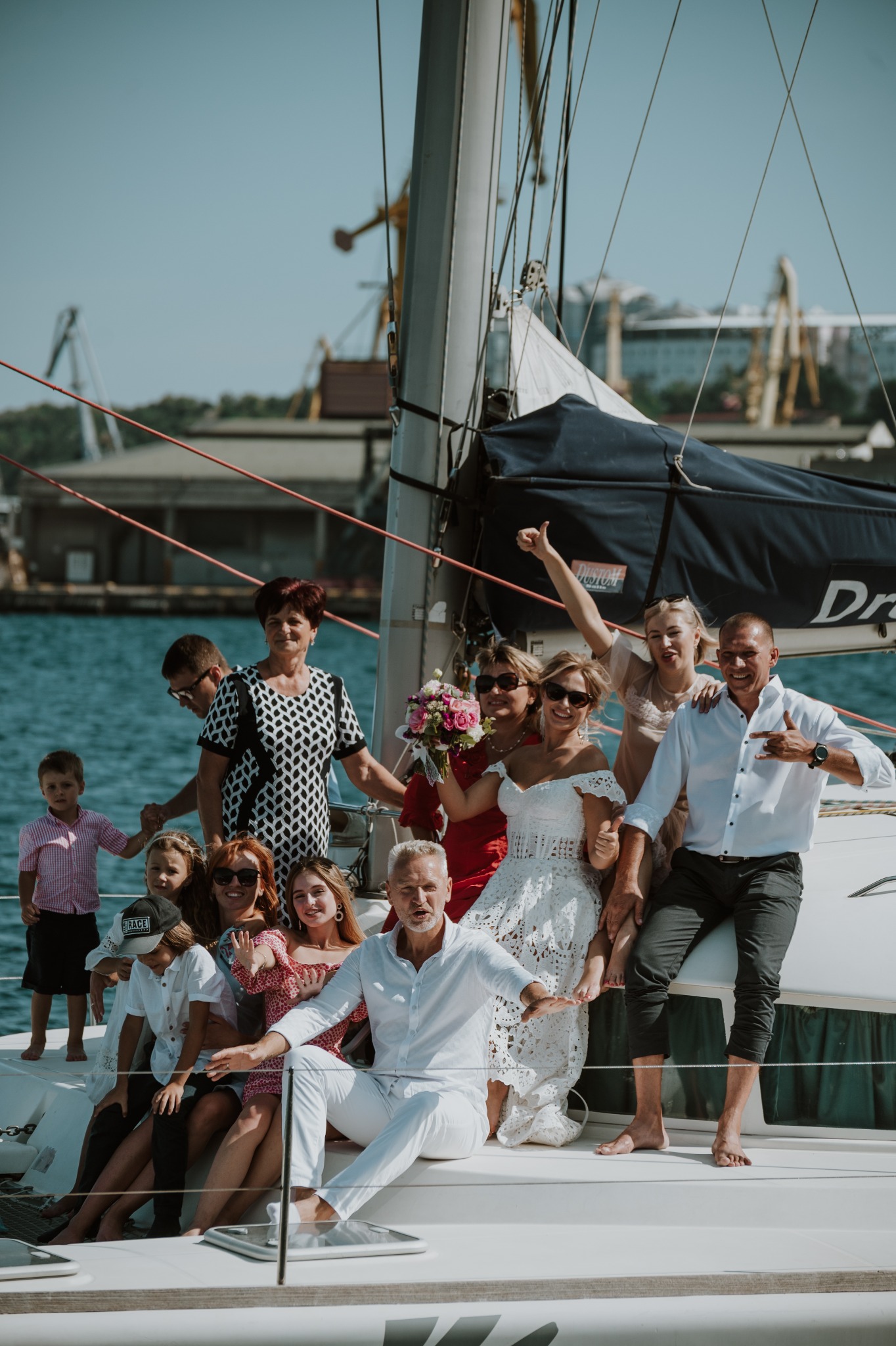 Duża grupa osób, w tym panna młoda w koronkowej sukni, pozuje na jachcie w porcie, z dźwigami widocznymi w tle.