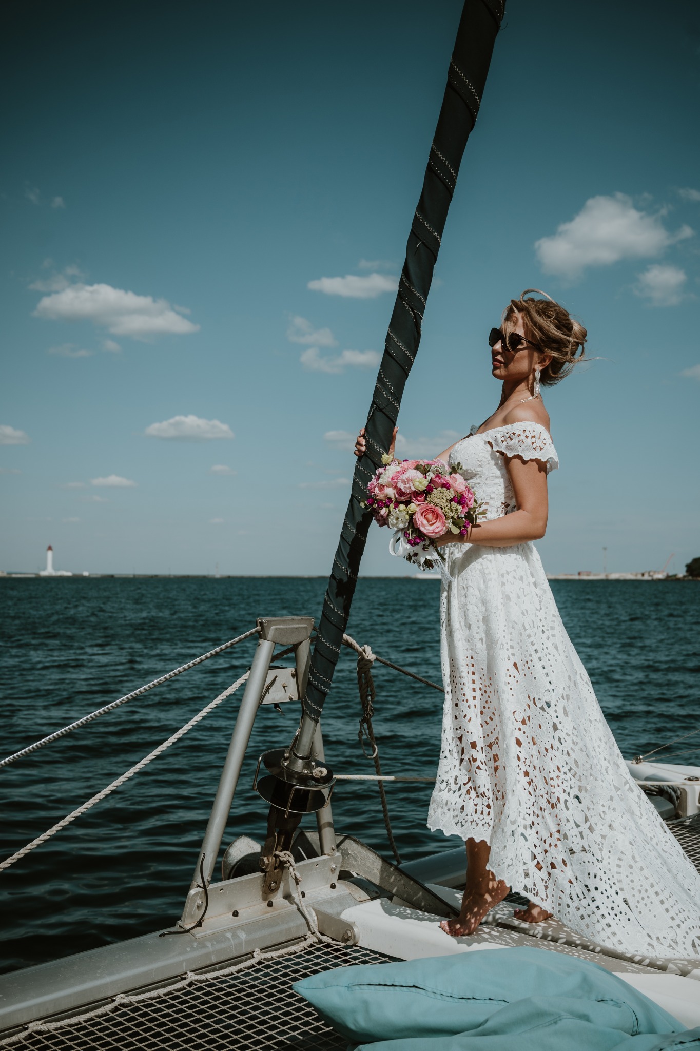 Panna młoda w koronkowej sukni i okularach przeciwsłonecznych, trzymająca bukiet różowych i białych kwiatów, pozuje na jachcie na tle morza i latarni morskiej.