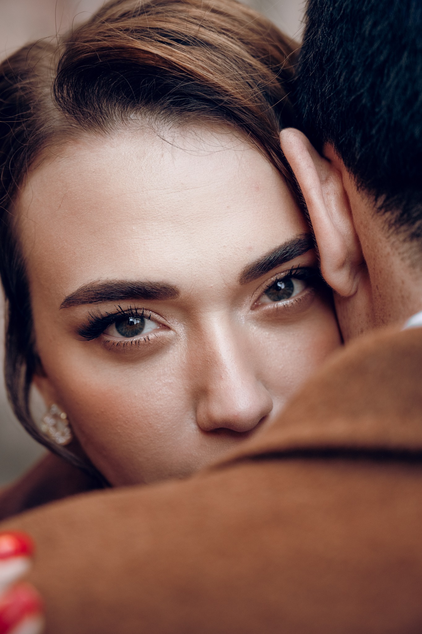 Portret kobiety z bliska, wtulona w mężczyznę w brązowym płaszczu, z widocznym kolczykiem i intensywnym spojrzeniem w obiektyw.
