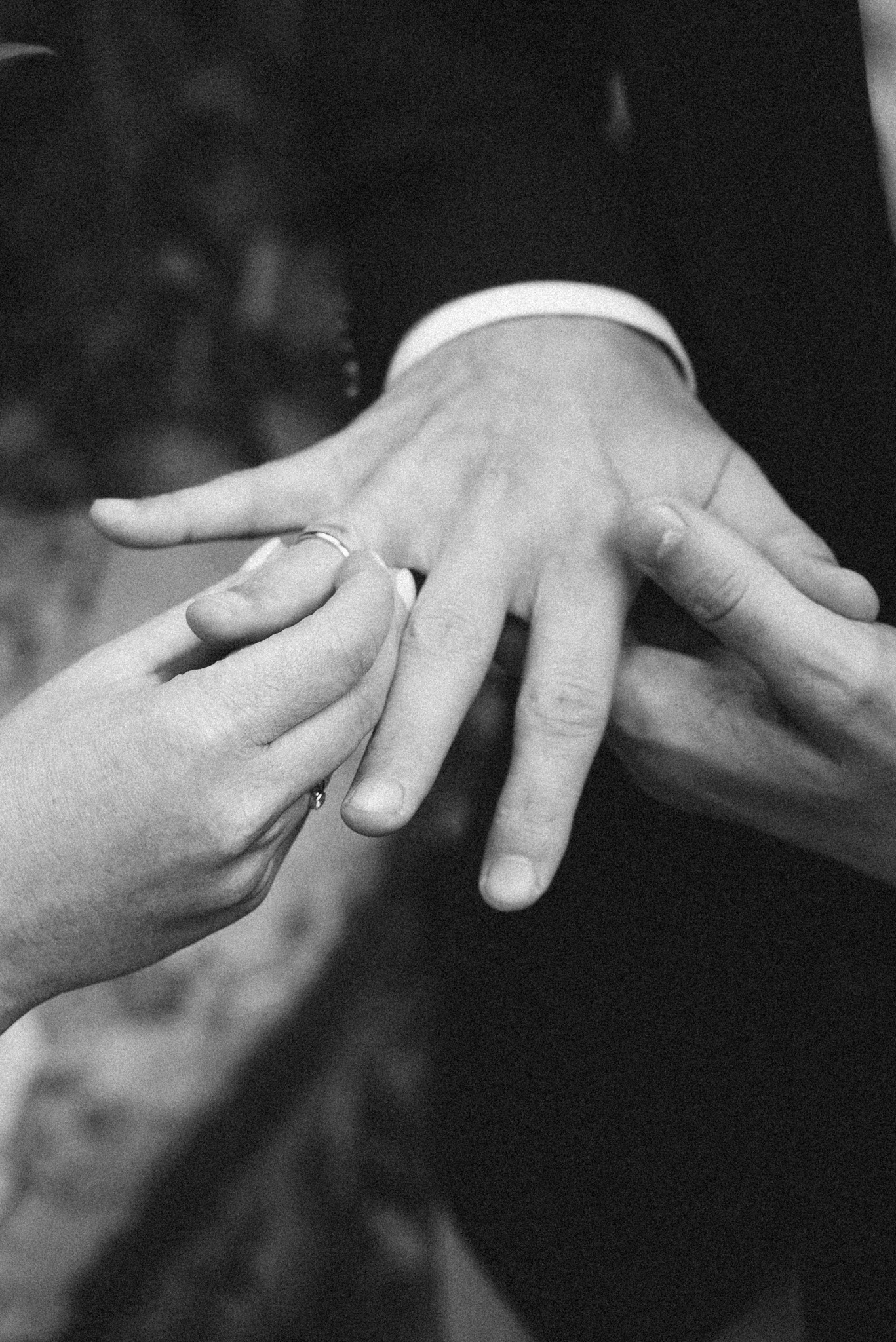 Czarno-białe zbliżenie dłoni zakładającej obrączkę na palec mężczyzny w garniturze.