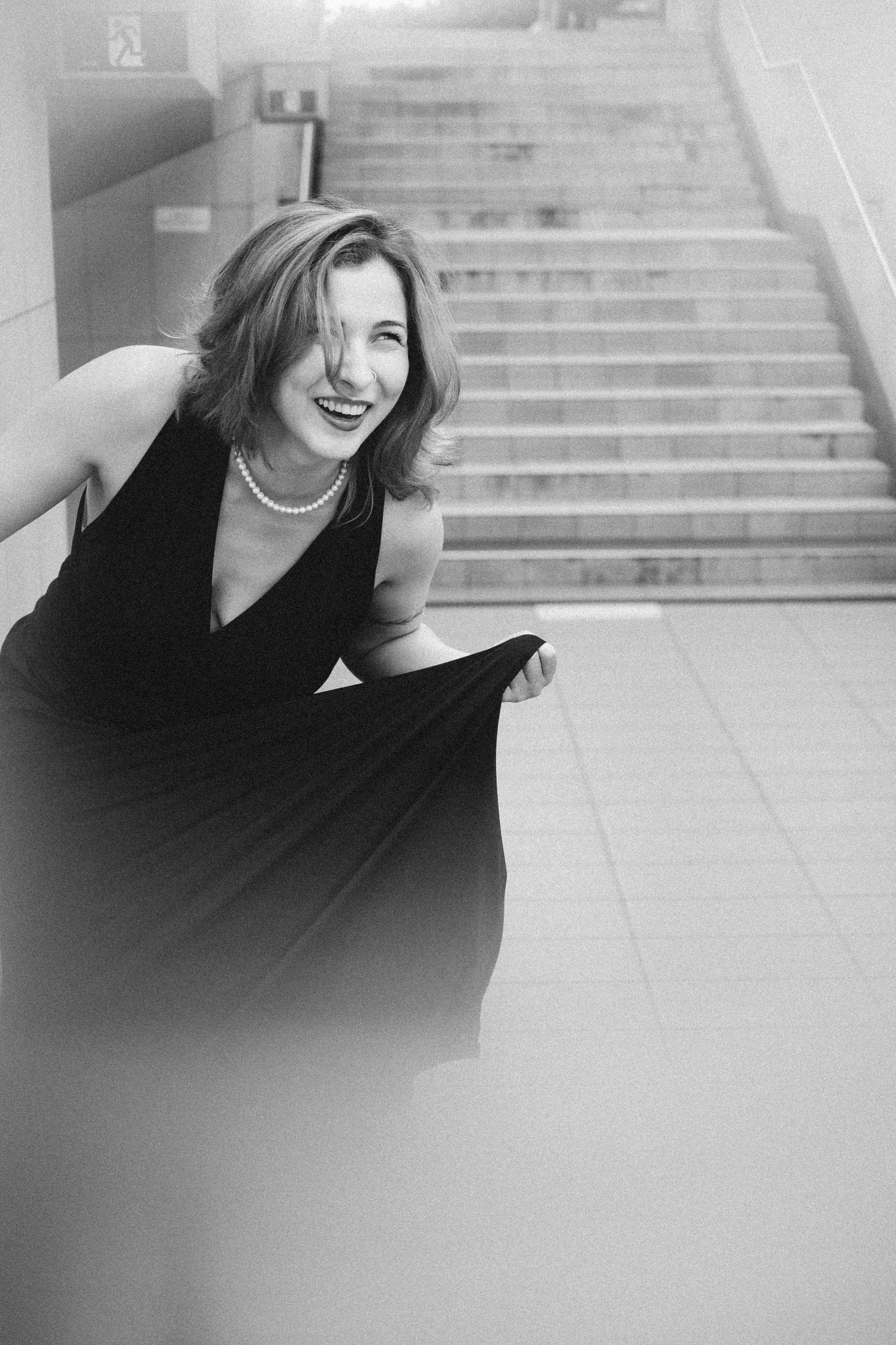 Uśmiechnięta kobieta w czarnej sukience i perłach, pozująca na tle betonowych schodów w czarno-białej sesji.