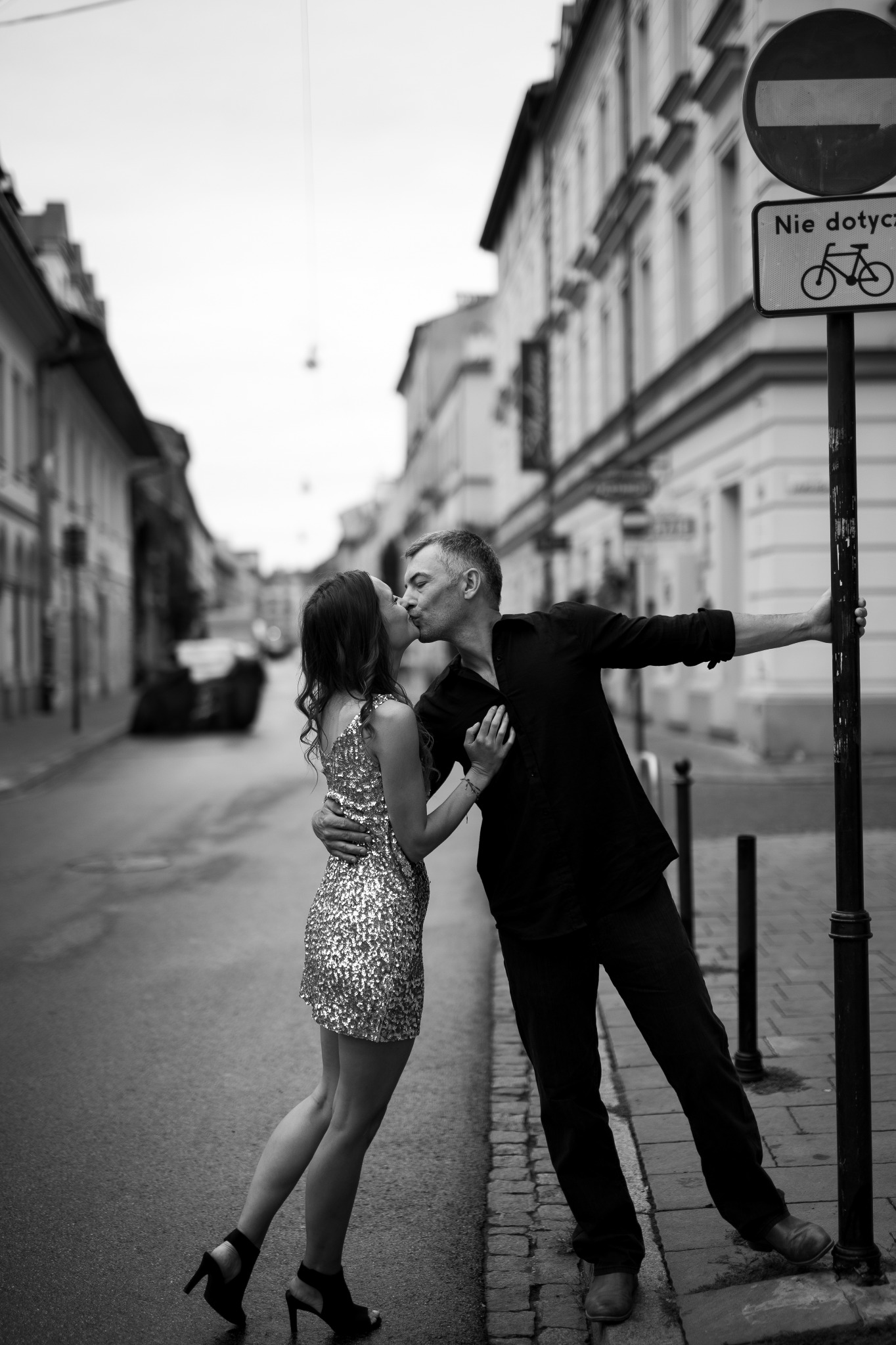 Czarno-białe zdjęcie pary całującej się na ulicy; kobieta w cekinowej sukience i szpilkach, mężczyzna oparty o znak drogowy, w tle budynki.