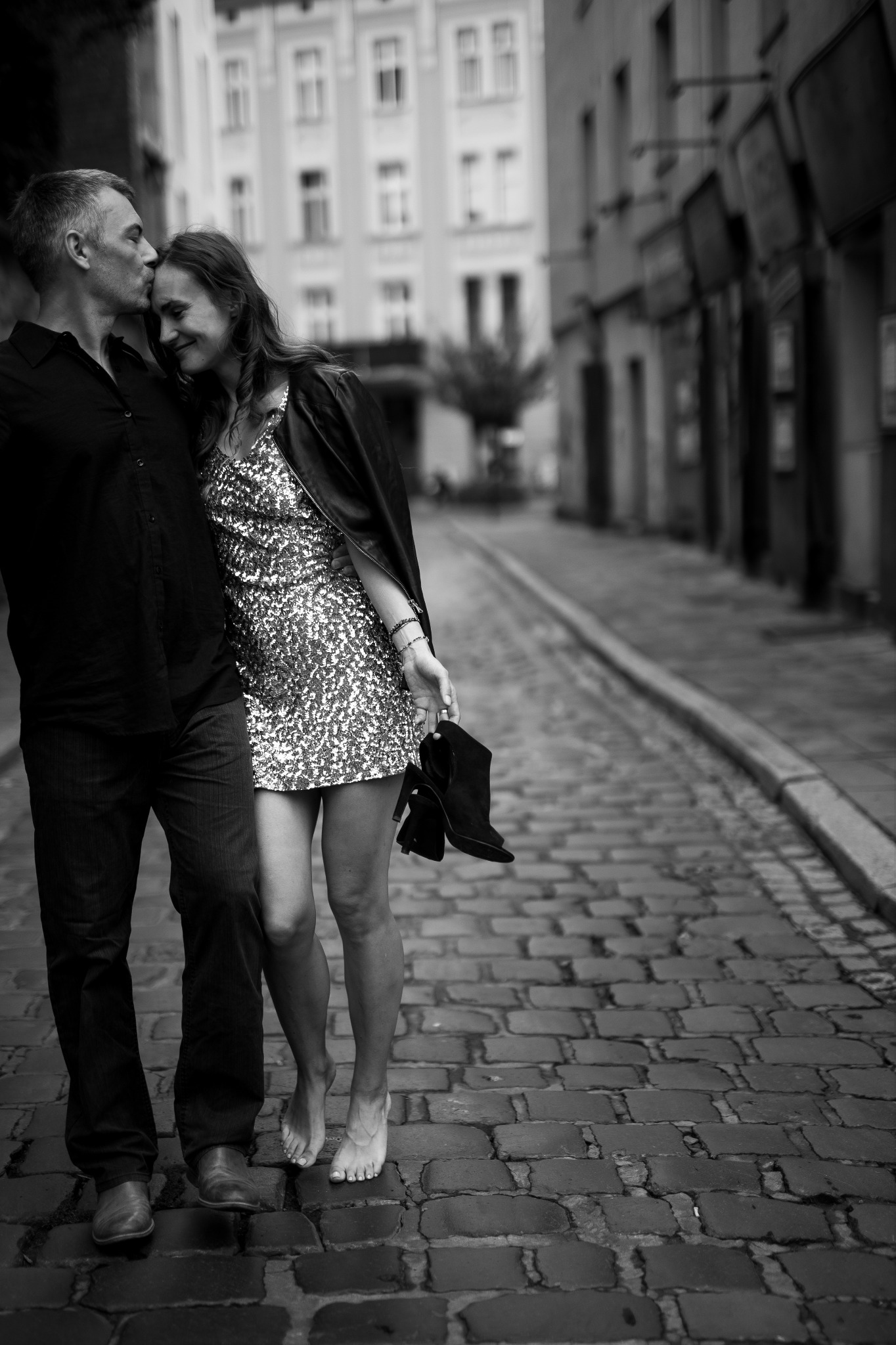 Czarno-białe zdjęcie pary na brukowanej uliczce: mężczyzna całuje kobietę w czoło, ona trzyma buty w ręce, boso, w cekinowej sukience i skórzanej kurtce.