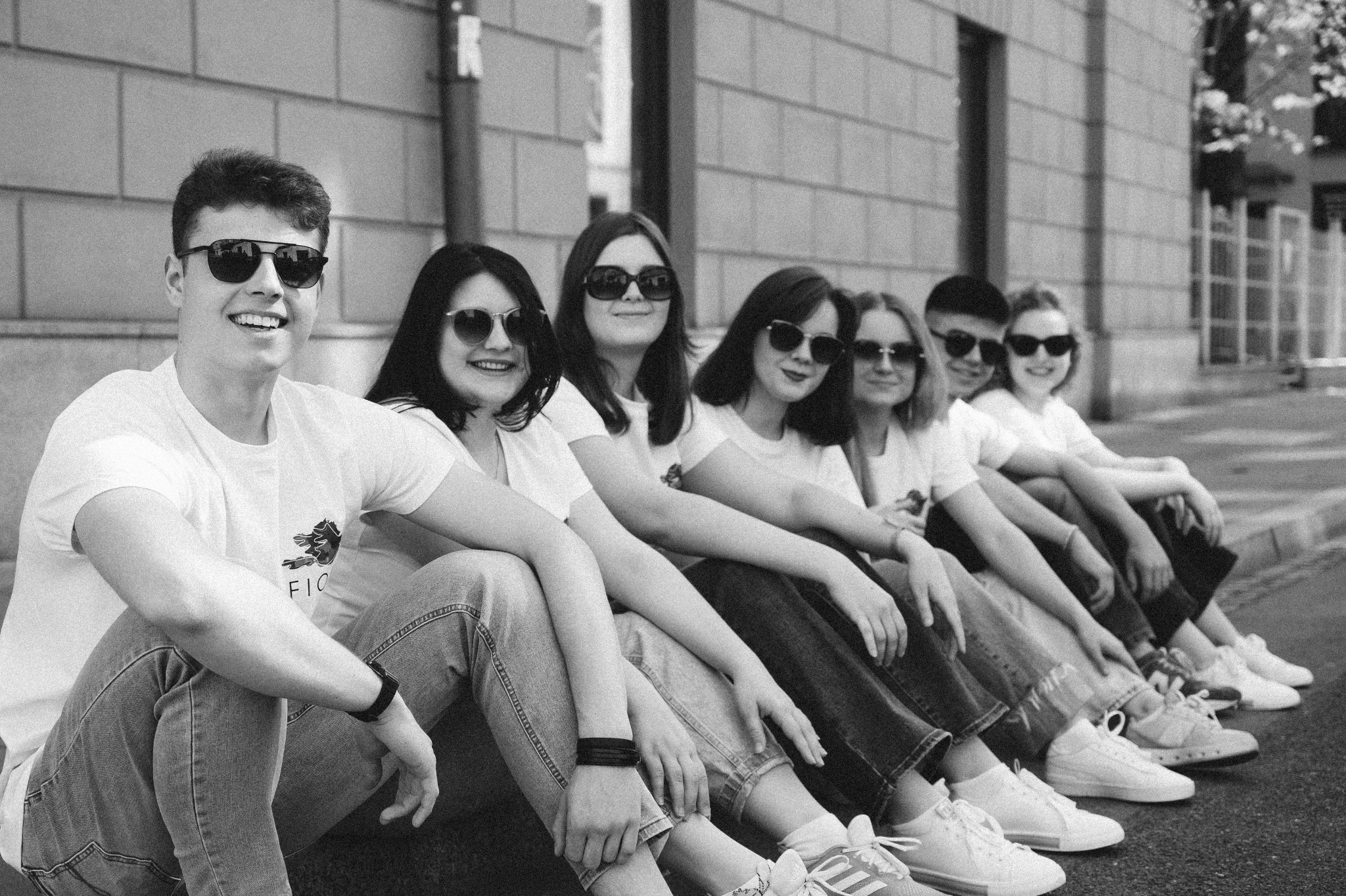 Czarno-białe zdjęcie grupy młodych osób siedzących rzędem na chodniku przed budynkiem, wszyscy w okularach przeciwsłonecznych, ubrani w jasne koszulki i jeansy, uśmiechnięci.
