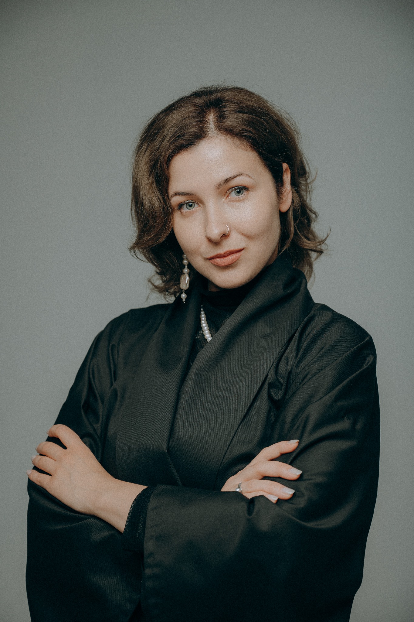 Portret kobiety z przekrzyżowanymi rękami, ubrana w czarną marynarkę, z kolczykiem w nosie i perłowymi kolczykami, na szarym tle.
